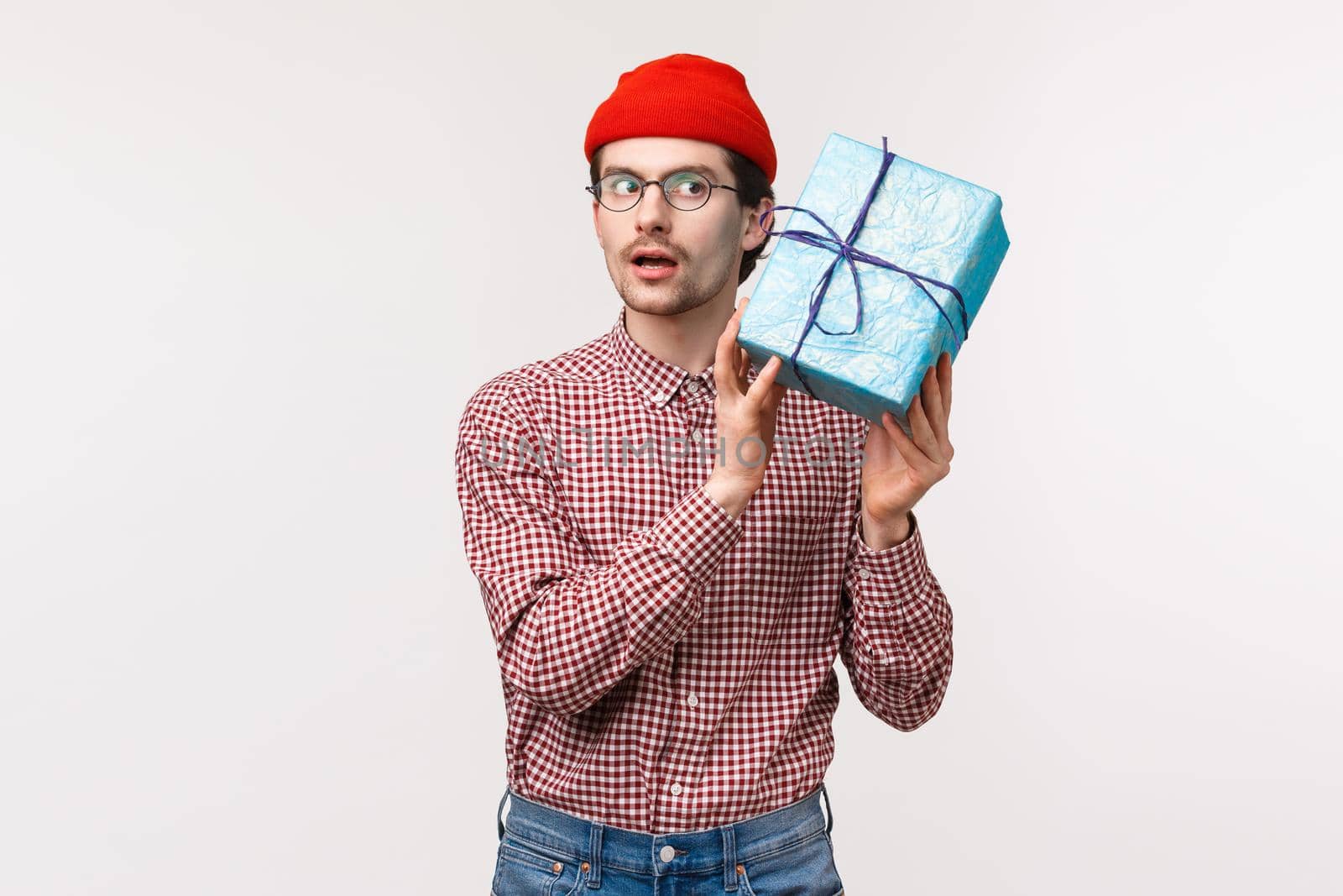 Waist-up portrait of curious funny caucasian bearded man in red beanie, glasses, shaking gift box near ear as trying guess whats inside, look focused, want to open surprise and see present.