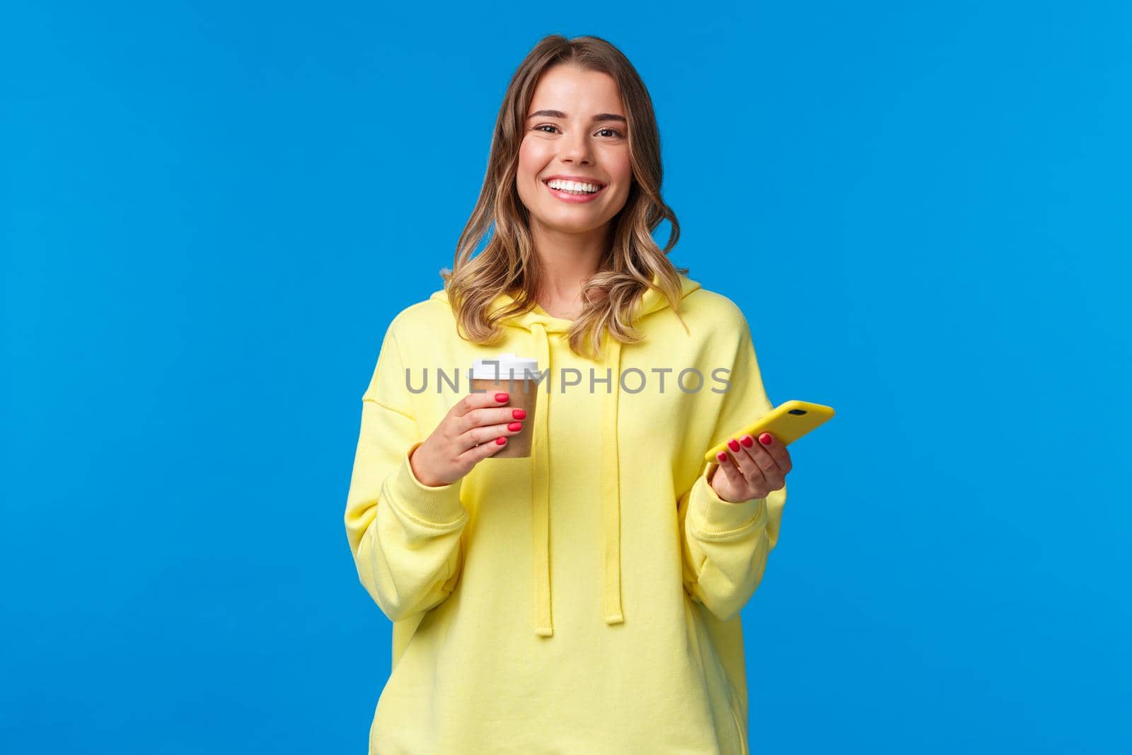 Cheerful young female student standing near cafe ordered take-away coffee, drinking from paper cup and holding mobile phone, look at camera with beaming white smile, satisfied by Benzoix