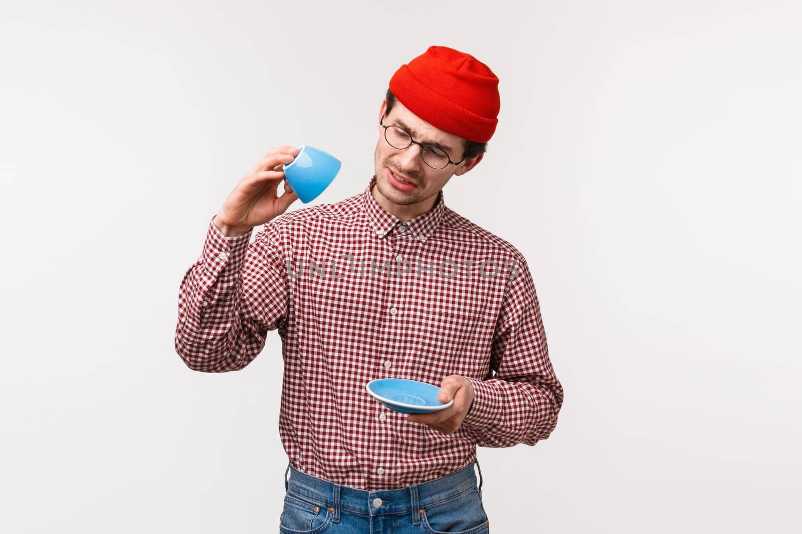 Waist-up portrait of funny european man with moustache in hipster beanie, glasses, look sad as no coffee left, look gloomy at empty cup, want drink this delicious tea some more, white background.
