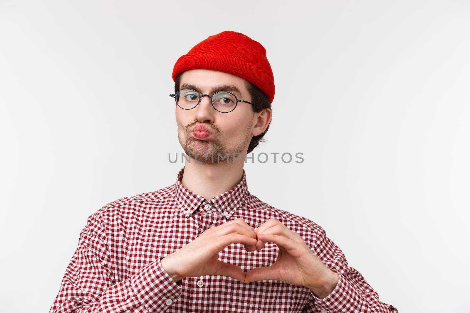 Close-up portrait funny caucasian male with geeky look, wear glasses and red beanie, pouting for kiss, showing heart gesture as confessing love, expressing sympathy in cringy way, white background.