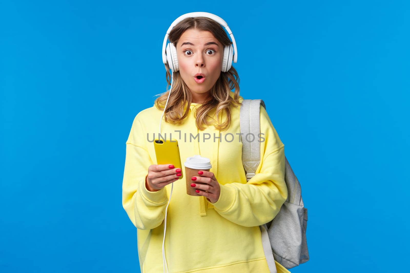 Surprised and amused young blond pretty girl look amazed camera say wow, wearing headphones and texting friend via smartphone, hold mobile phone, backpack and take-away coffee.