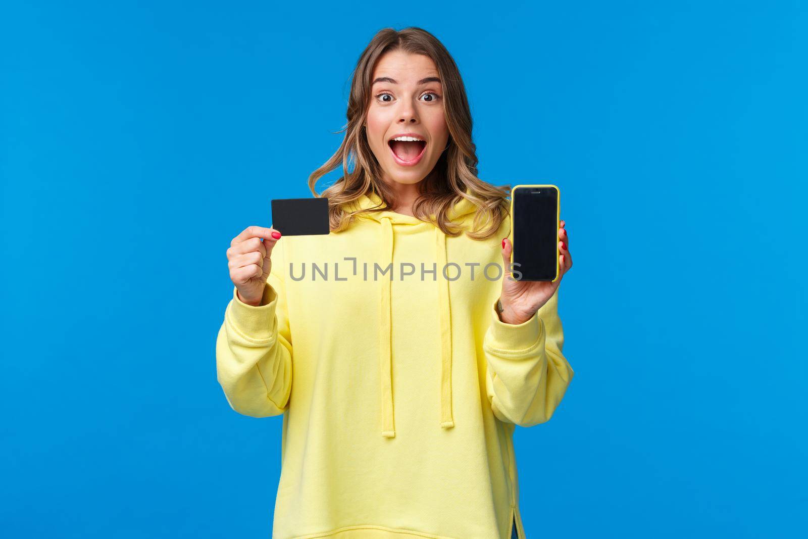 Amused and excited blond girl recommend using banking service, showing smartphone display and credit card and looking camera fascinated as receive bonus to bank account, blue background.