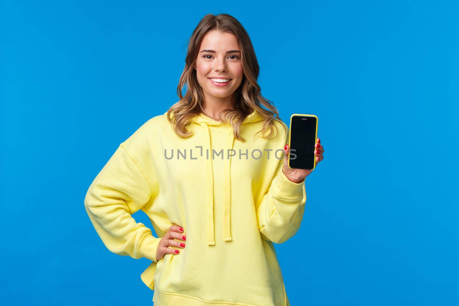 Waist-up portrait good-looking blond girl in yellow hoodie promote smartphone application, show mobile phone display and smiling camera as recommend subscribe or download app.