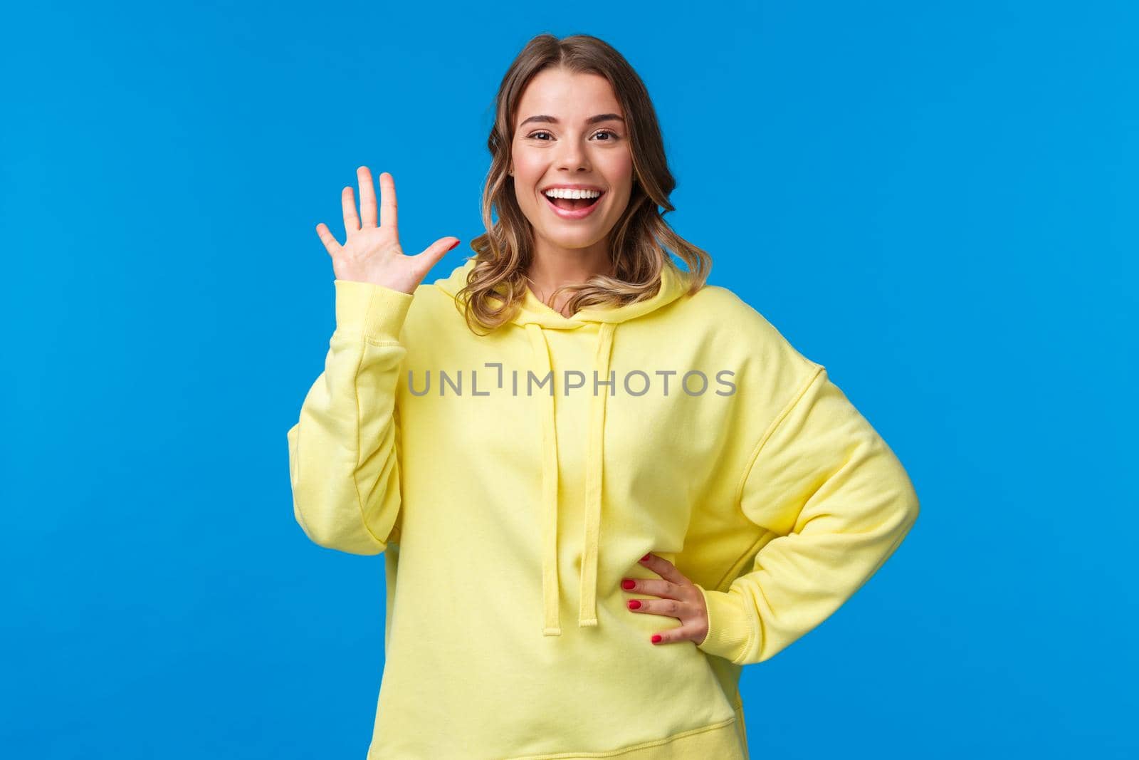 Cheerful friendly outgoing blond girl waving hand, informal greeting concept, smiling white teeth saying hi, welcoming new members to company, see friend, stand blue background.