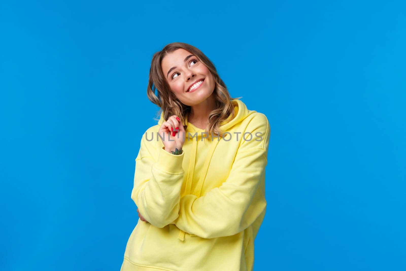 Dreamy silly young caucasian woman with blond hair, looking up thoughtful fantasizing, imaging something with curious smile, thinking or dreaming, standing blue background romantic.