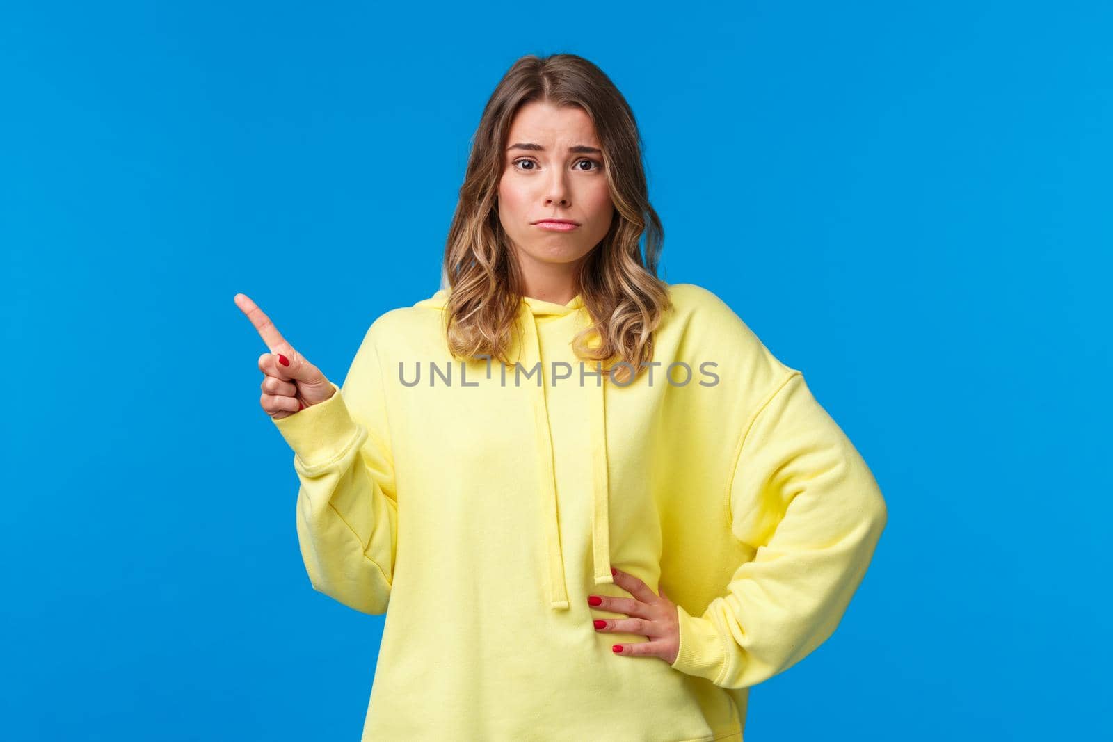 Upset and insecure cute european blond young woman in yellow hoodie, having doubts, being skeptical about topic, pointing finger upper left corner and look unsure camera, blue background.