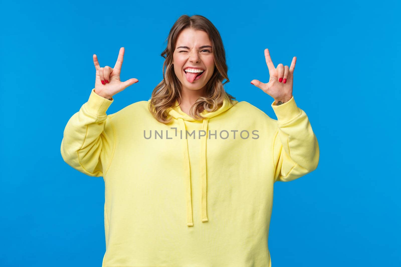 Let us have fun. Carefree joyful good-looking blonde female in yellow hoodie show rock-n-roll sign and tongue as feel excited and pleased with awesome party, dancing enjoy concert.