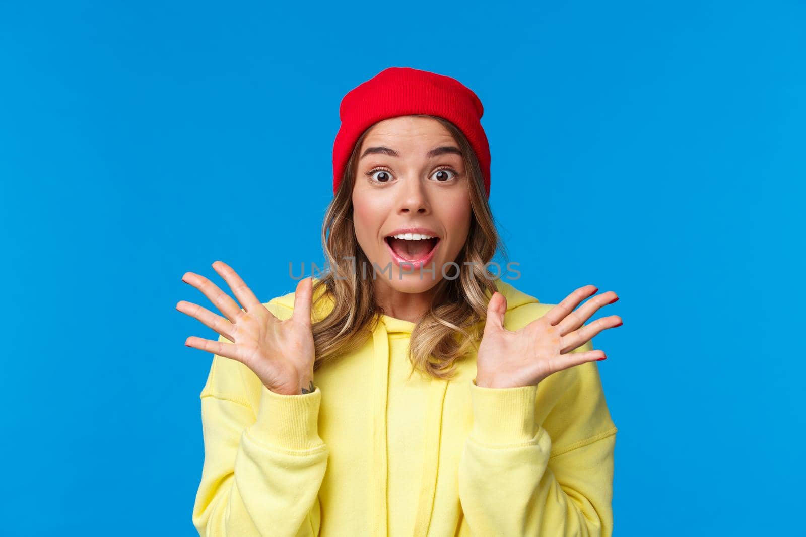 Excited good-looking european female explain great news, talking to friend astonished, shaking hands thrilled and looking camera fascinated, standing blue background in red beanie.