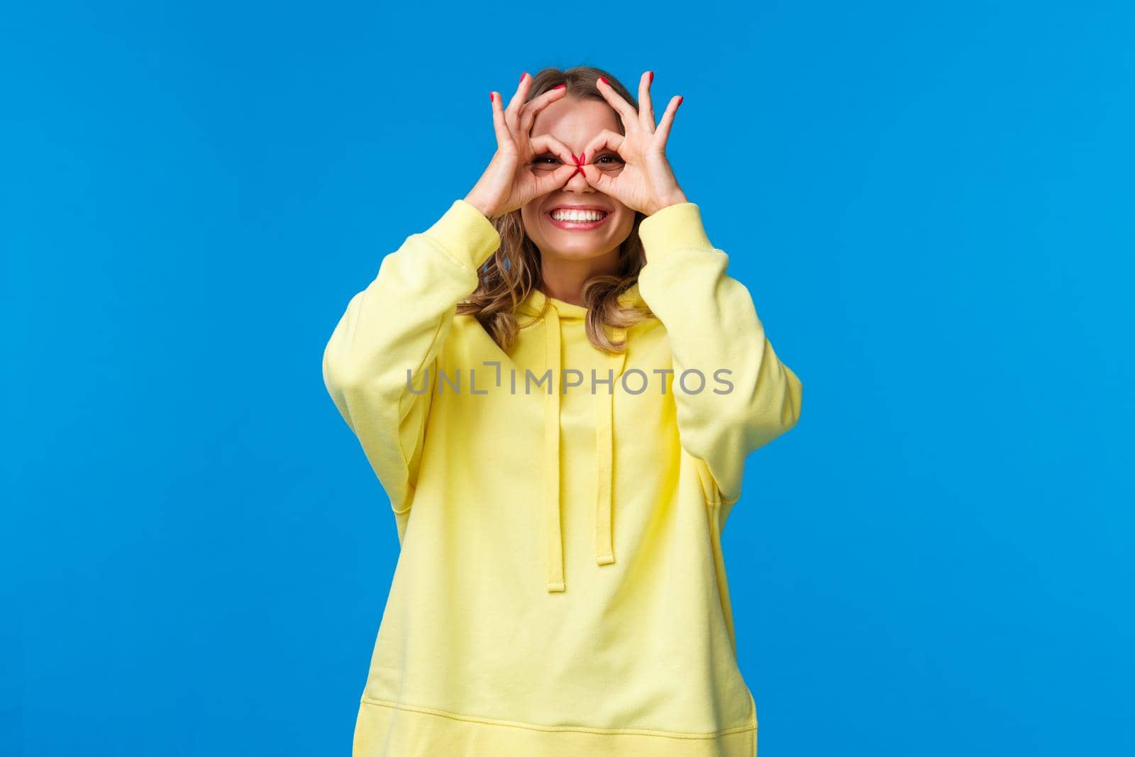 Dreamy and silly girl looking through fingers with happy playful expression, make okay gestures form glasses with hands and smiling carefree, look far away, searching something, blue background by Benzoix