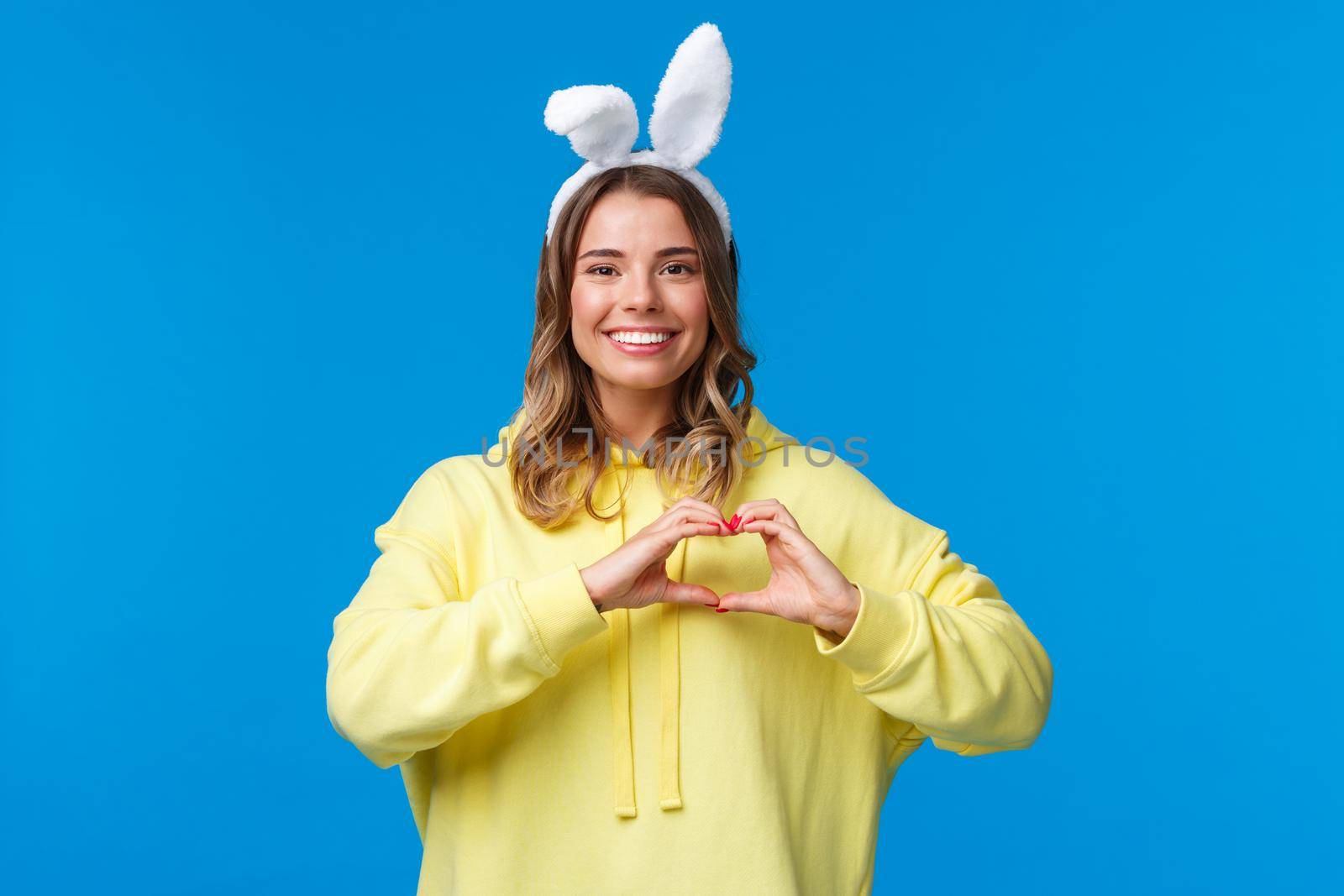 Holidays, traditions and celebration concept. Waist-up portrait of charismatic cute blond girl in rabbit ears, congratulate everyone with Easter holiday, show heart gesture and smiling.