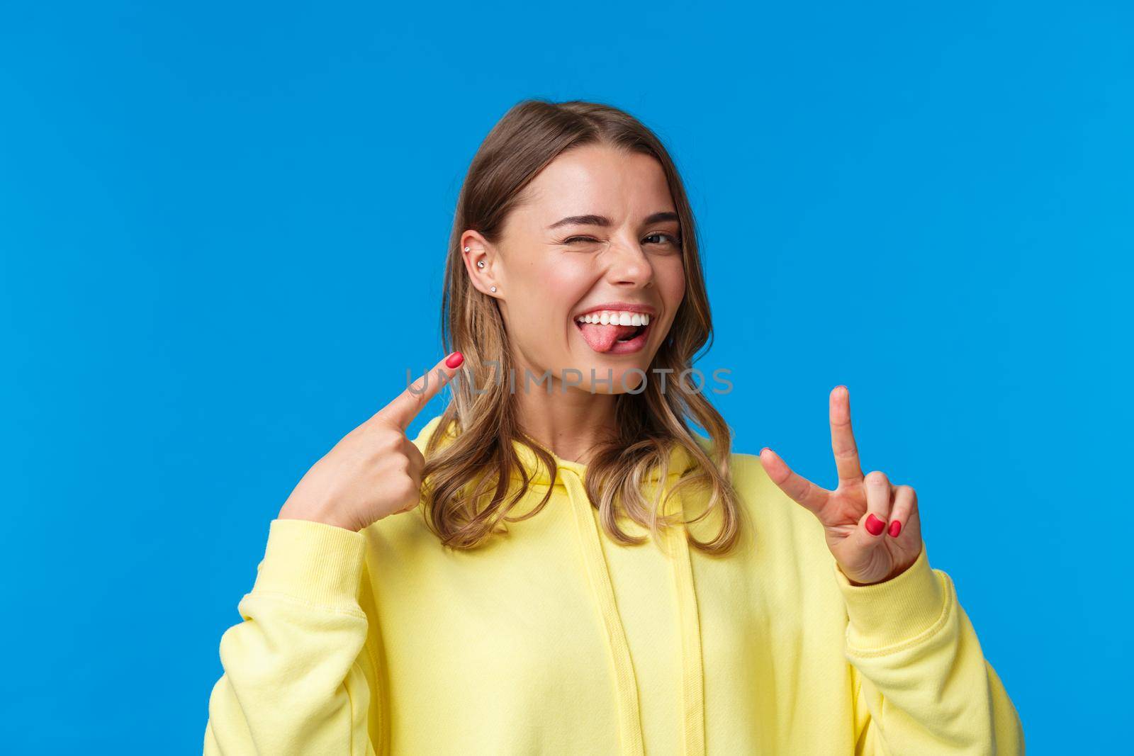 Close-up portrait of carefree enthusiastic blond girl in yellow hoodie, with pierced ear, show tongue and wink joyful, pointing herself make peace gesture, standing blue background.