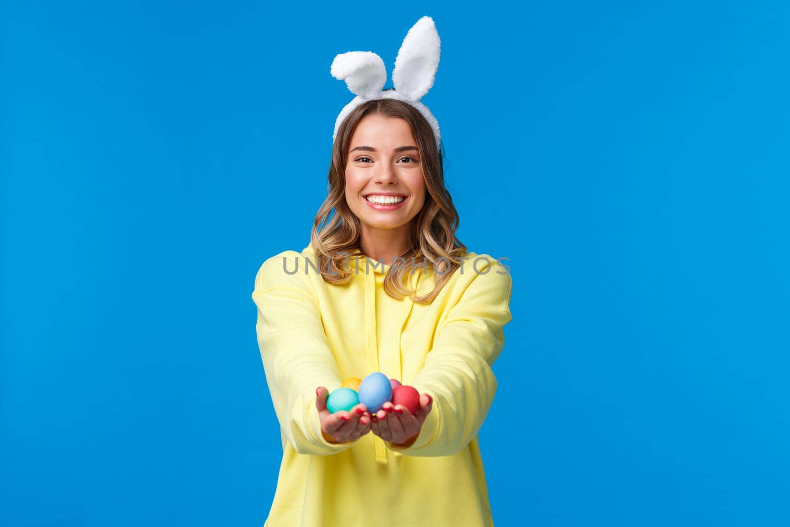 Holidays, people and fun concept. Charismatic cute blond girl holding coloured Easter eggs, wearing bunny ears and smiling joyfully, congratulate family with holy day, blue background.