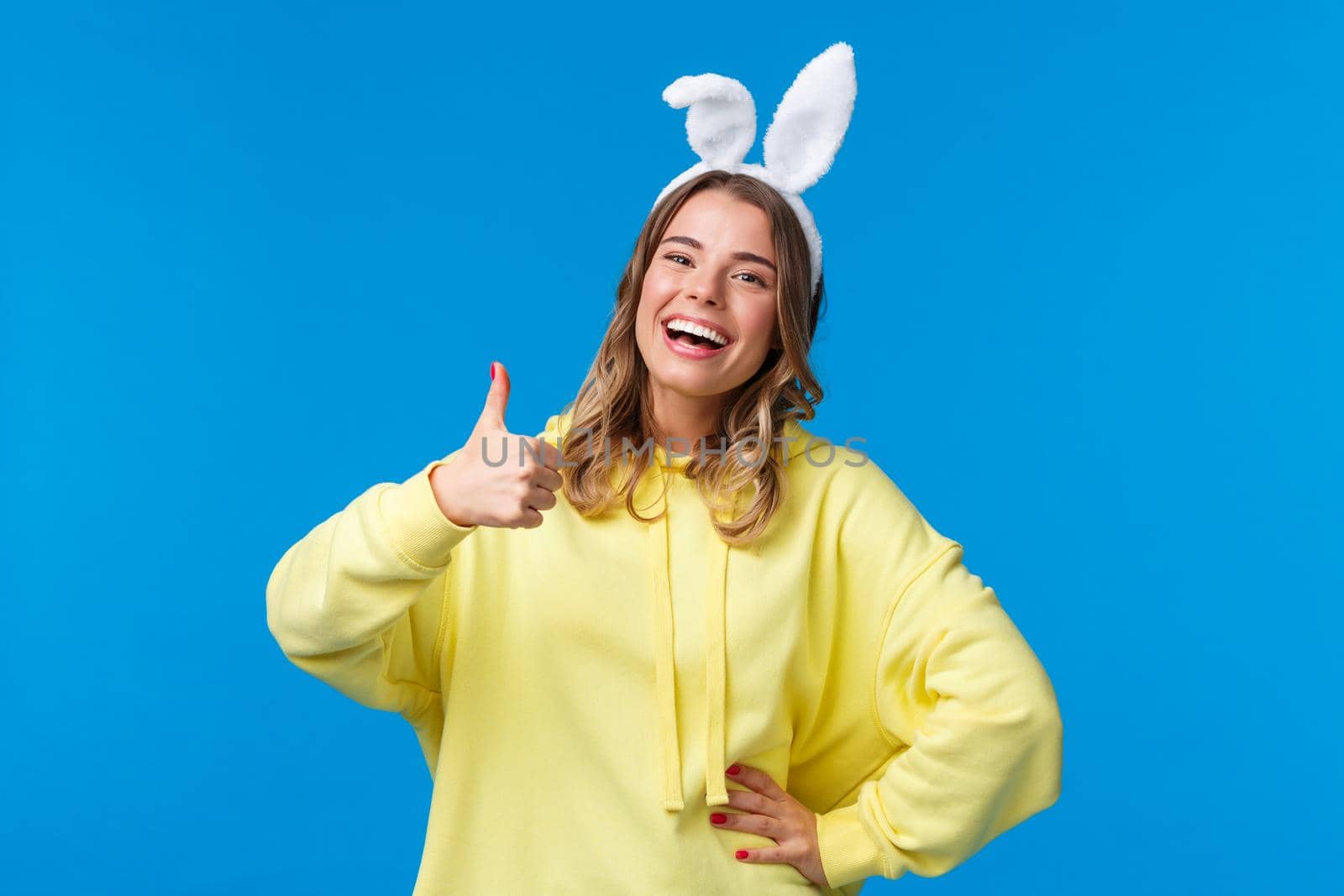 Holidays, traditions and celebration concept. Optimistic cute blond girl with rabbit ears laughing and showing approval gesture, thumb-up praise nice work, happy Easter, blue background.