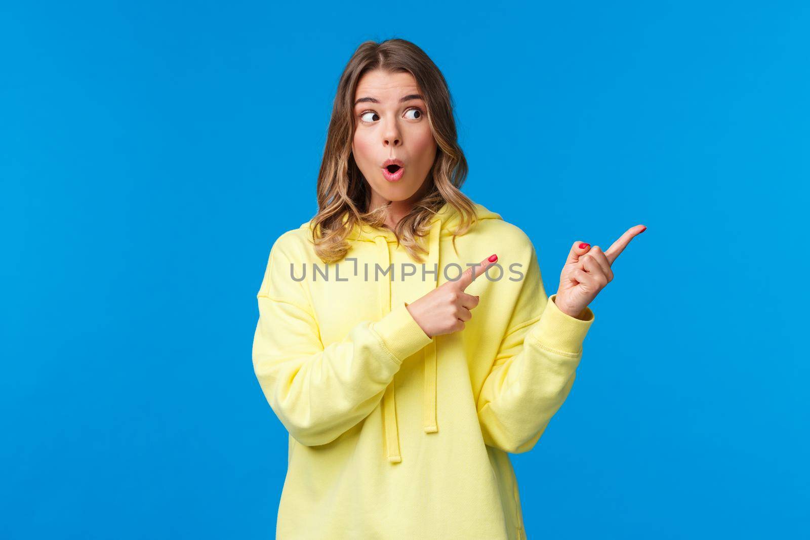 Wow something interesting. Intrigued and surprised alluring blond woman in yellow hoodie talking about last rumors, pointing and looking right with impressed expression, blue background by Benzoix