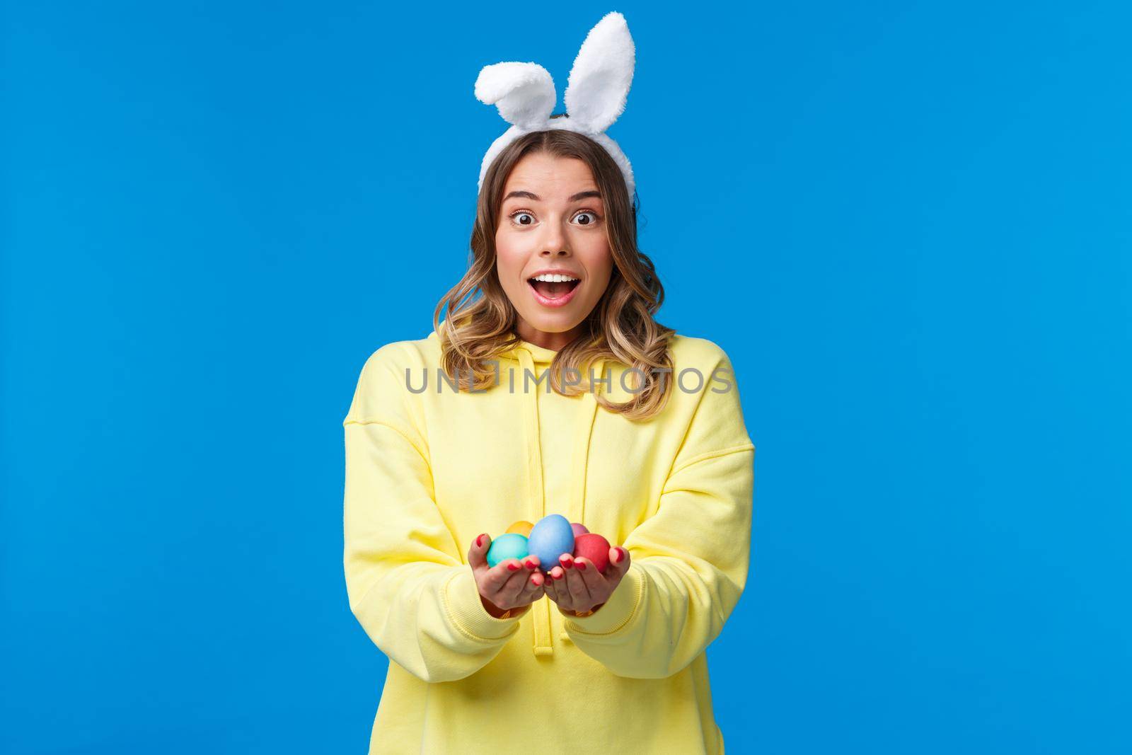 Amused cute and lovely caucasian blond woman in rabbit ears holding Easter eggs, give it to family member as celebrating divine holiday together, standing blue background by Benzoix