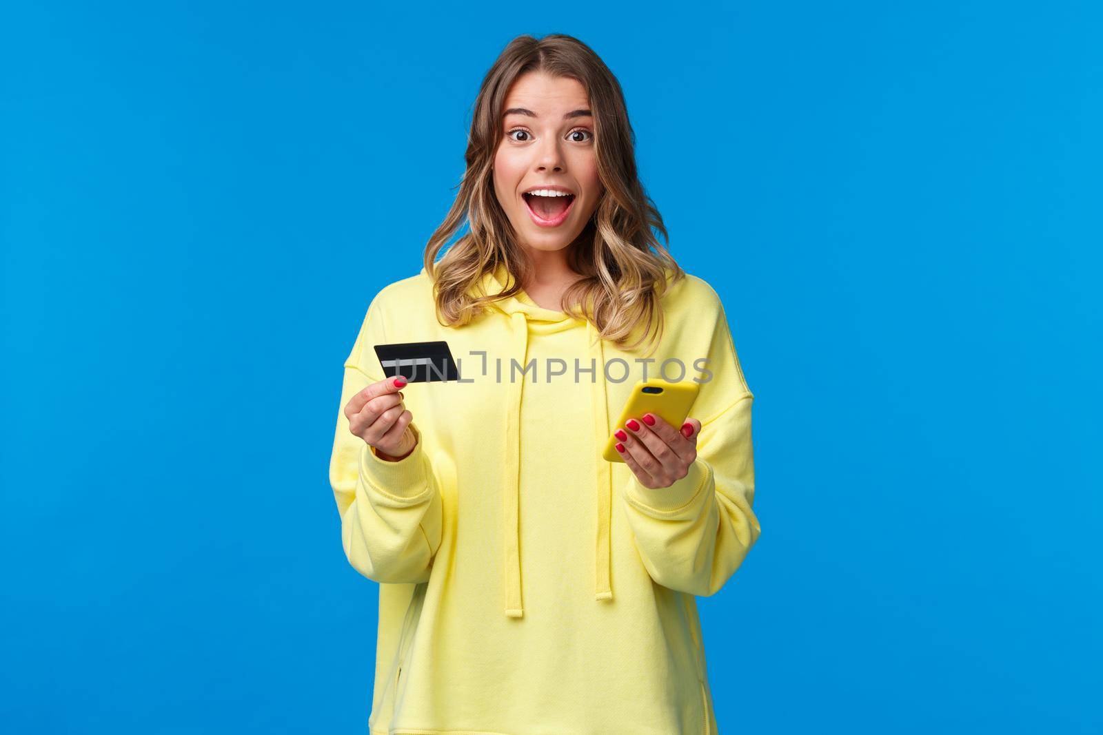 Amused and surprised cute blond girl receive cool cashback or banking offer after using new credit card with special student offer, holding mobile phone and smiling camera.