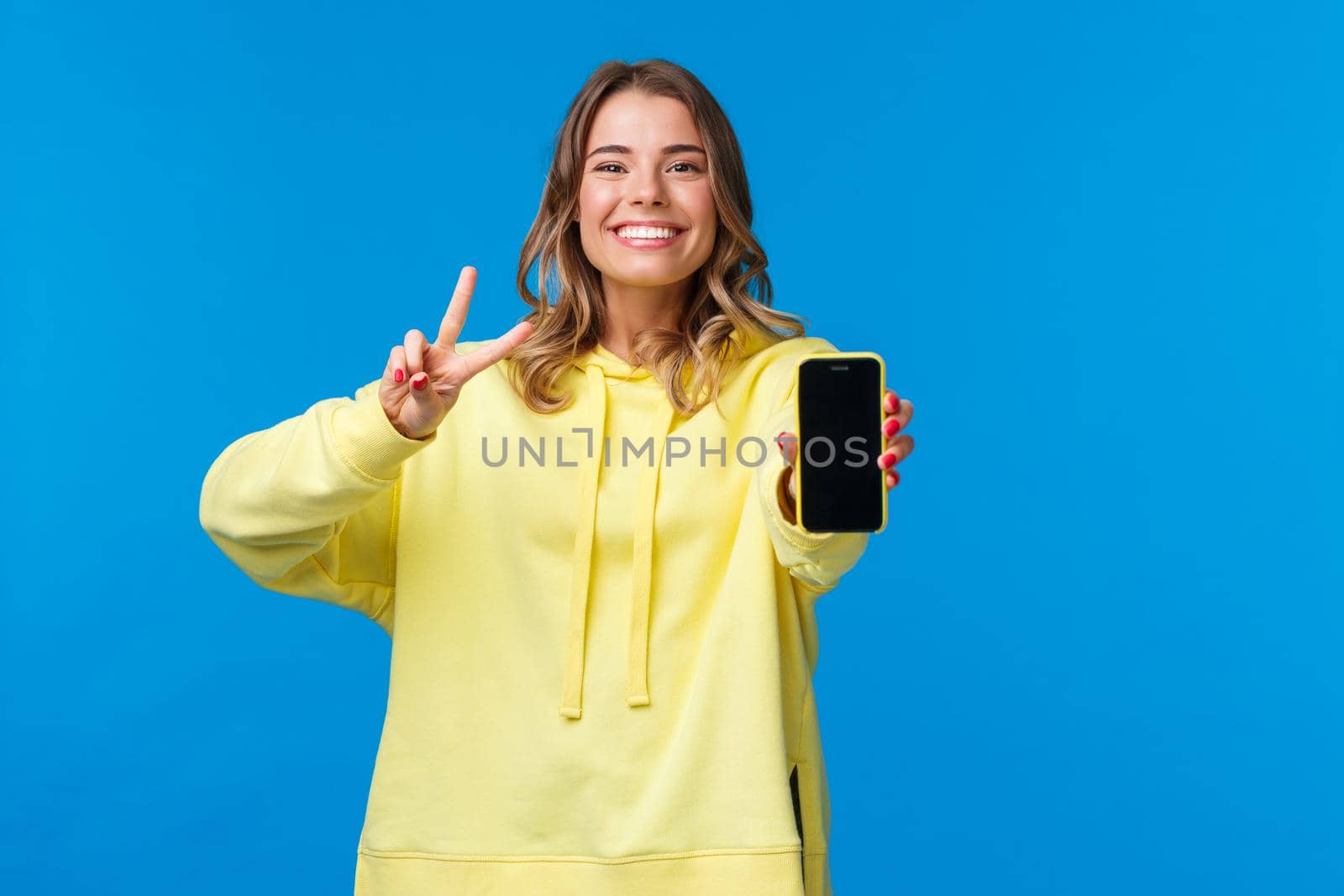 Cute smiling happy blond girl in yellow hoodie showing you her online blog or social media page on mobile phone display, make peace gesture and grinning, stand blue background by Benzoix