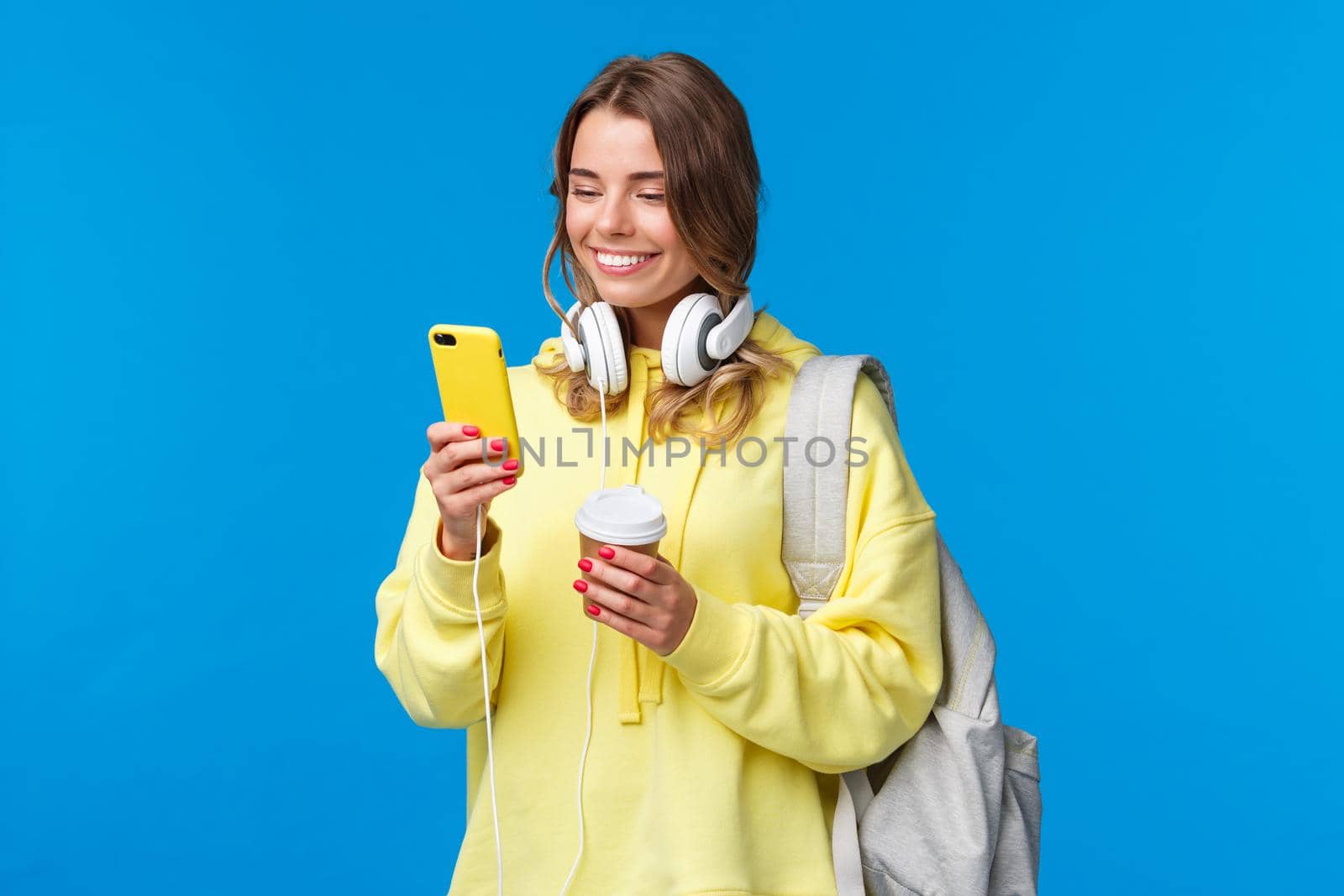 Girl using mobile phone to contact friend after college, texting as walking along street with backpack, take-away coffee cup and headphones, smiling joyfully smartphone display by Benzoix