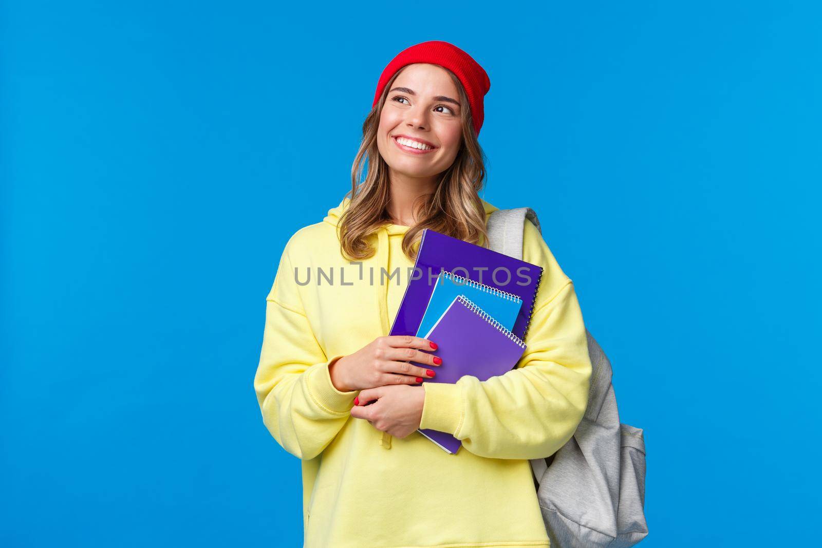 Dreamy cute smart college girl, female student in hipster red beanie, yellow hoodie, looking thoughtful upper left corner imaging summer holidays, smiling holding notebooks and backpack by Benzoix