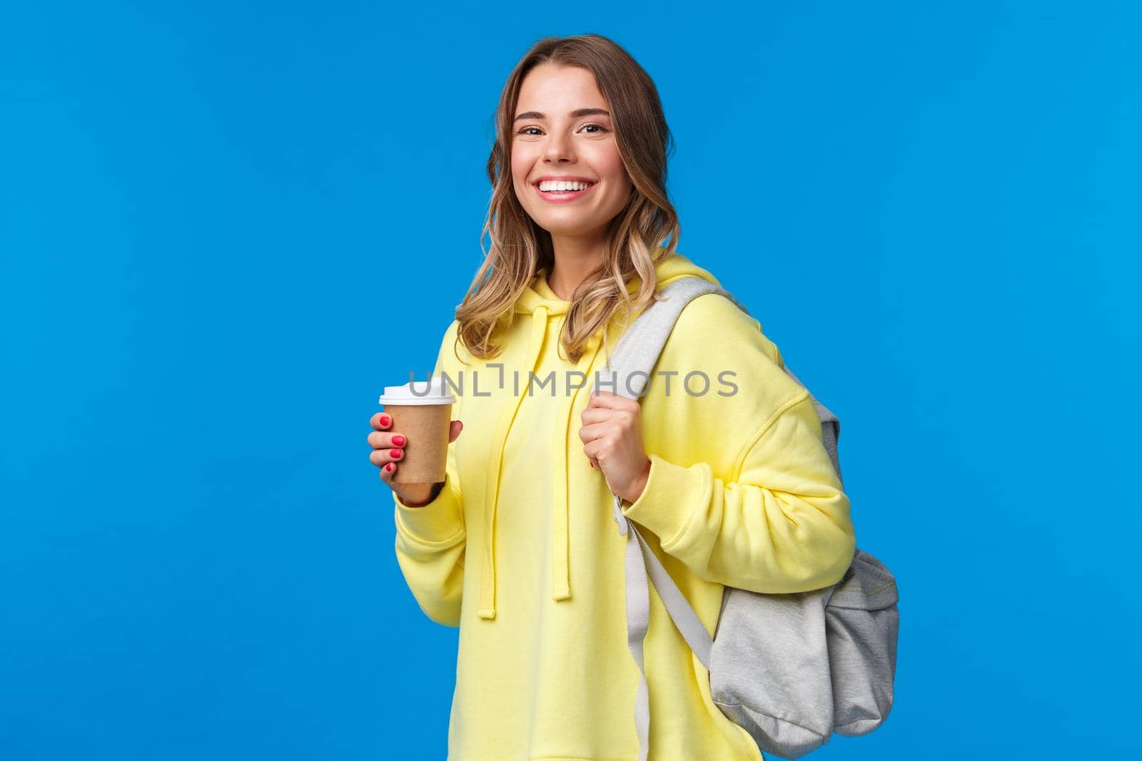 Joyful young blond female student with backpack smiling camera satisfied as holding cup of take-away coffee from favorite cafe after classes, drinking beverage, stand blue background by Benzoix