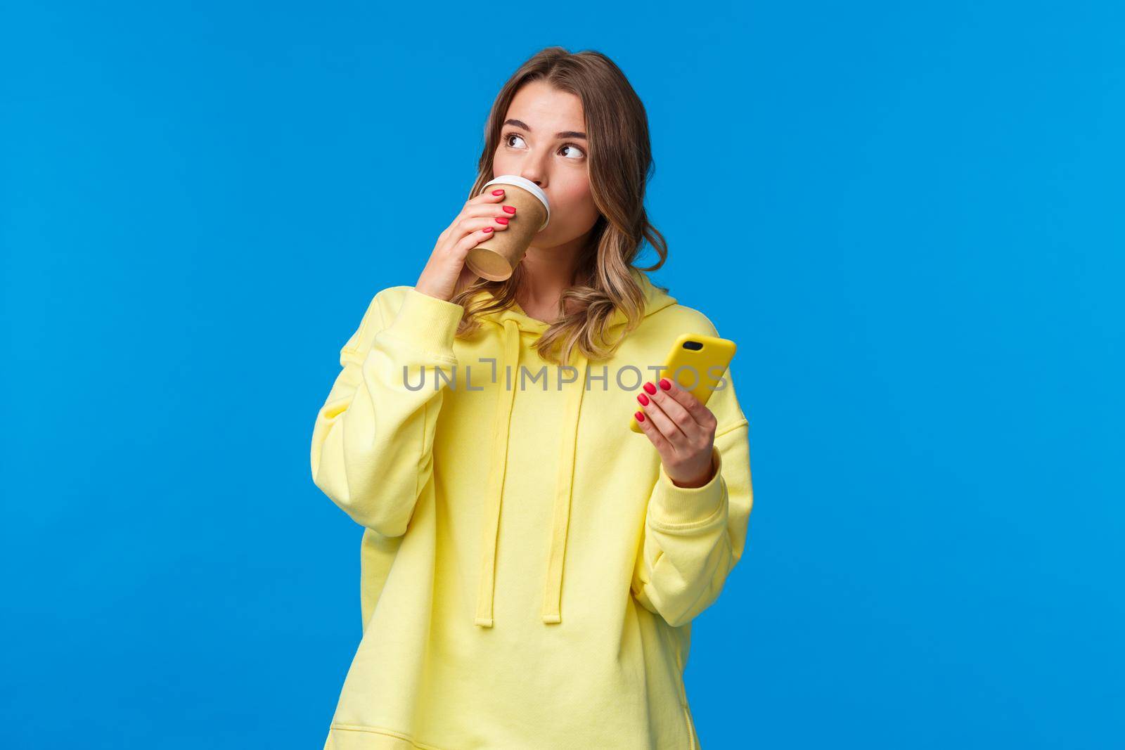 Thoughtful cute young blond female smm manager working freelance, drinking coffee from take-away cup look aside pondering and holding mobile phone, standing blue background by Benzoix