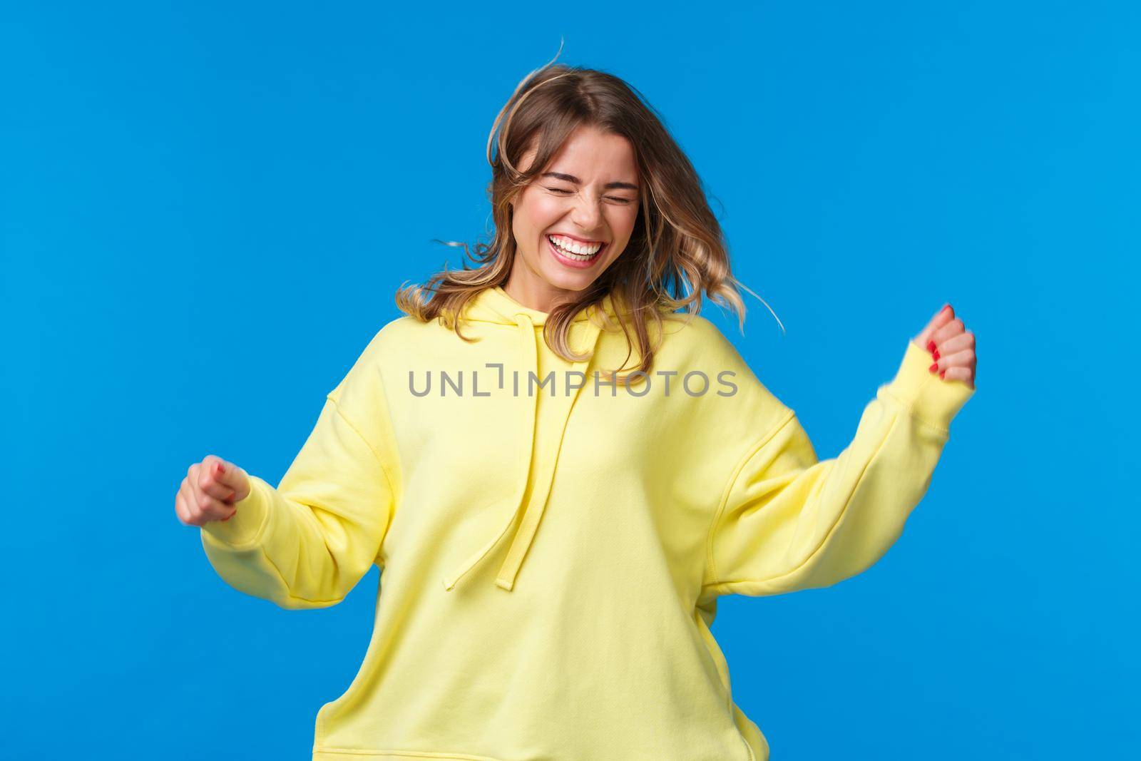 Girls just want ahve fun. Carefree and happy laughing blond girl enjoy spring, jumping and dancing as listening music on concert, shaking head as hair floating in air, blue background.