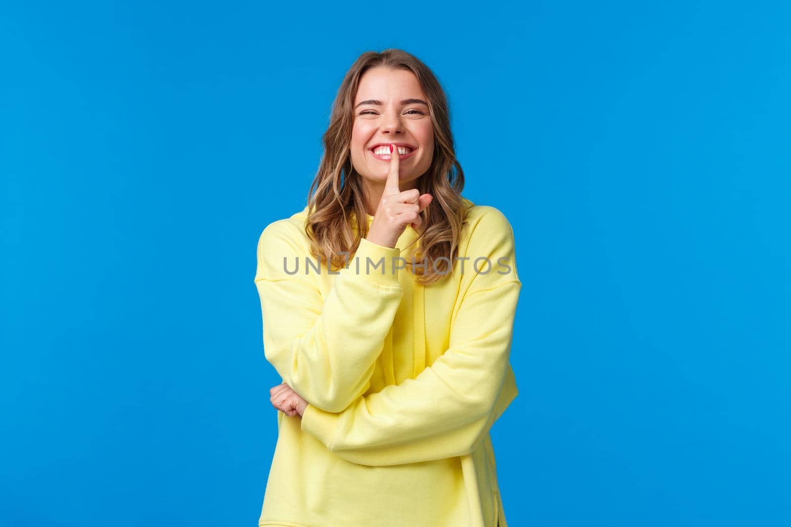 Happy cheerful silly blond caucasian woman in yellow hoodie hiding secret, prepare b-day surprise for boyfriend, shush with index finger pressed to lips as smiling joyfully, blue background by Benzoix