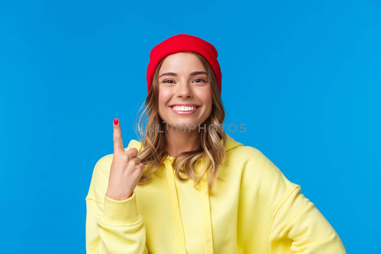 Close-up portrait of cute optimistic european female in red beanie and yellow hoodie, showing number one, order single person table in cafe, smiling happy, standing blue background.