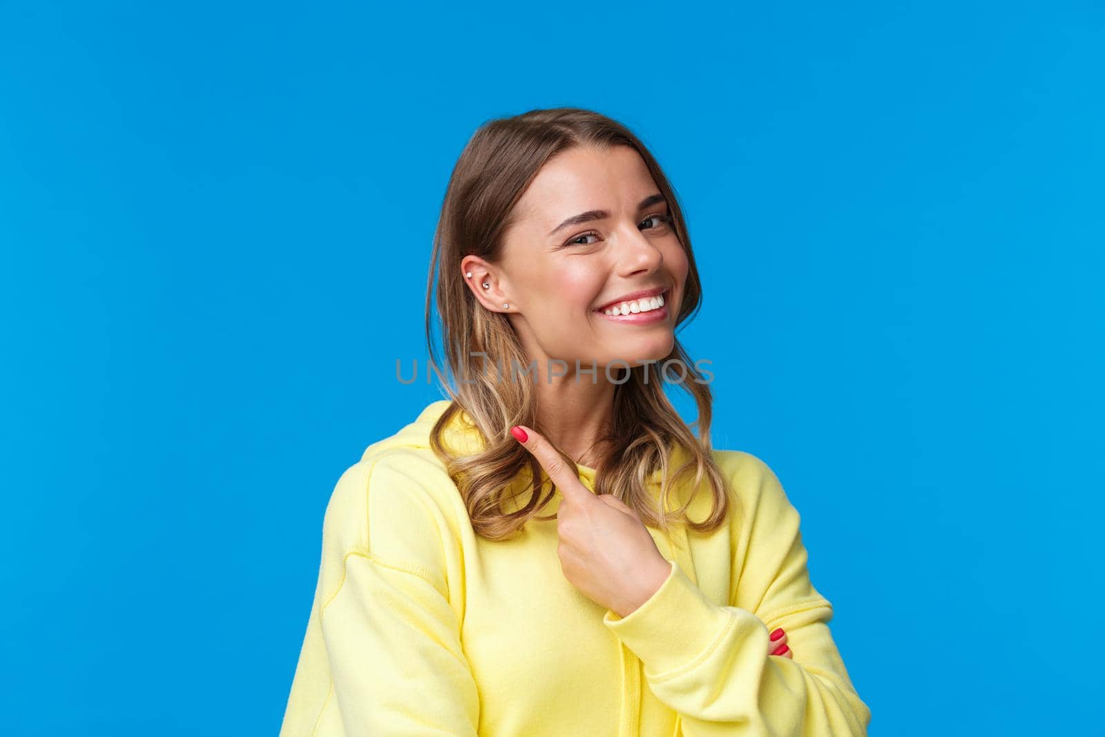 Close-up portrait of beautiful blond girl with short hair pointing at her piercing in ear, smiling proud and satisfied, change her look, standing blue background in yellow hoodie. Copy space