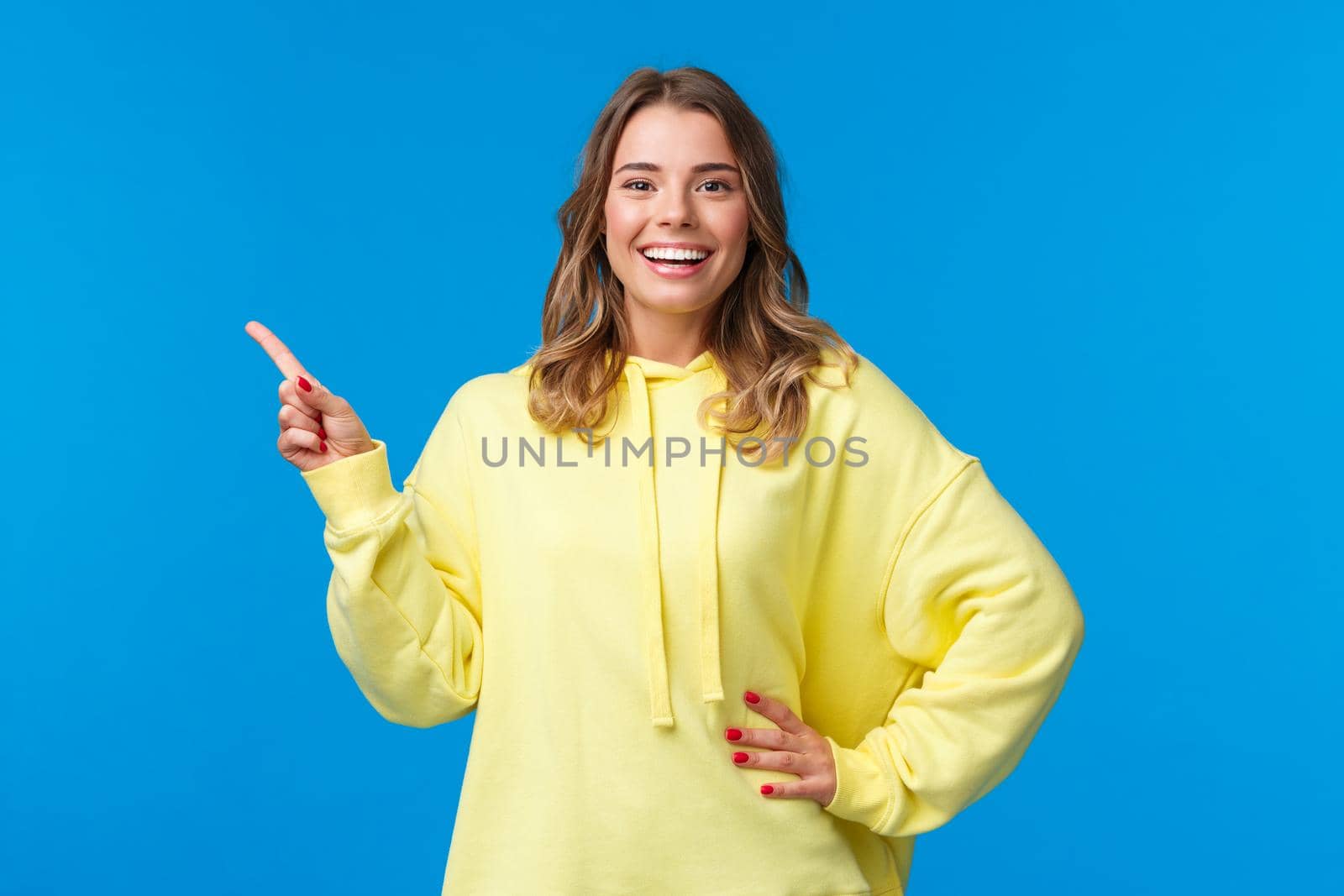 Take a look. Upbeat good-looking blond girl pointing finger left, recommend promo, advertise product, look pleased and smiling camera, explain project, standing blue background by Benzoix