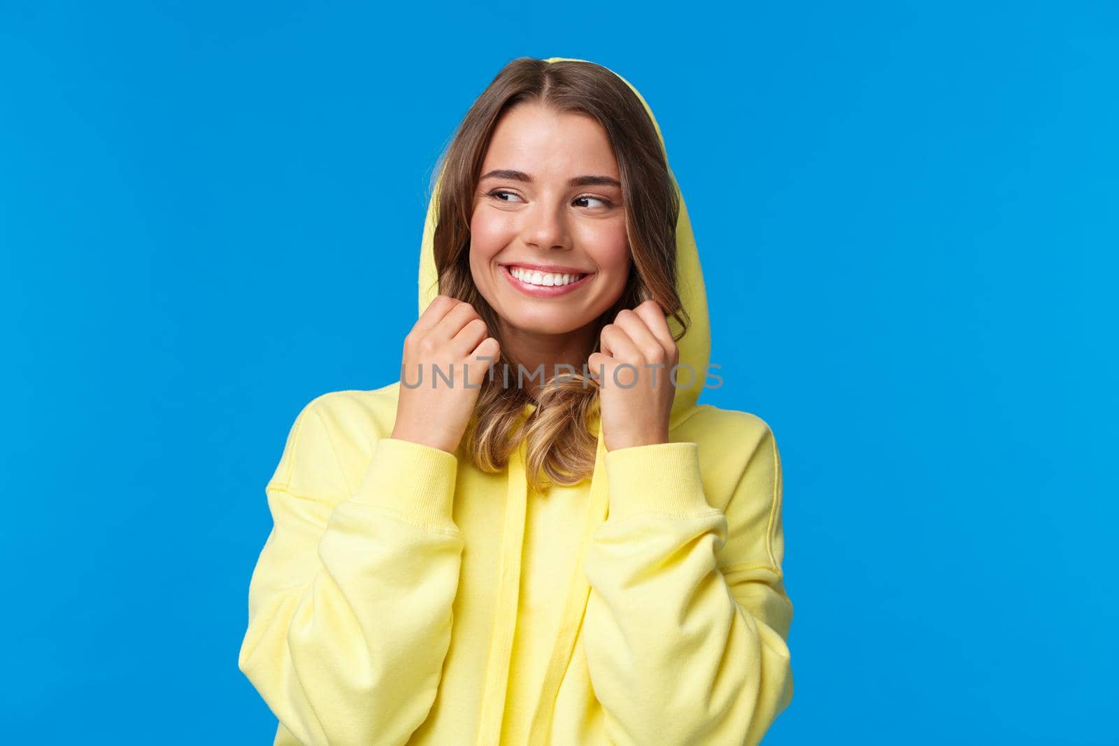 Close-up portrait beautiful charming blond girl put on hood of sweatshirt and smiling, look away, feel chilly as walking on spring day, standing carefree over blue background. Copy space