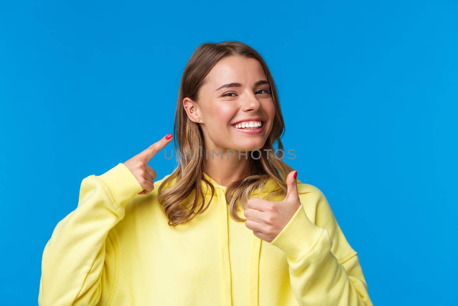 Cheerful and pleased, satisfied good-looking european female pointing at pierced ear and smiling delighted, show thumbs-up like her new jewelry, standing blue background by Benzoix