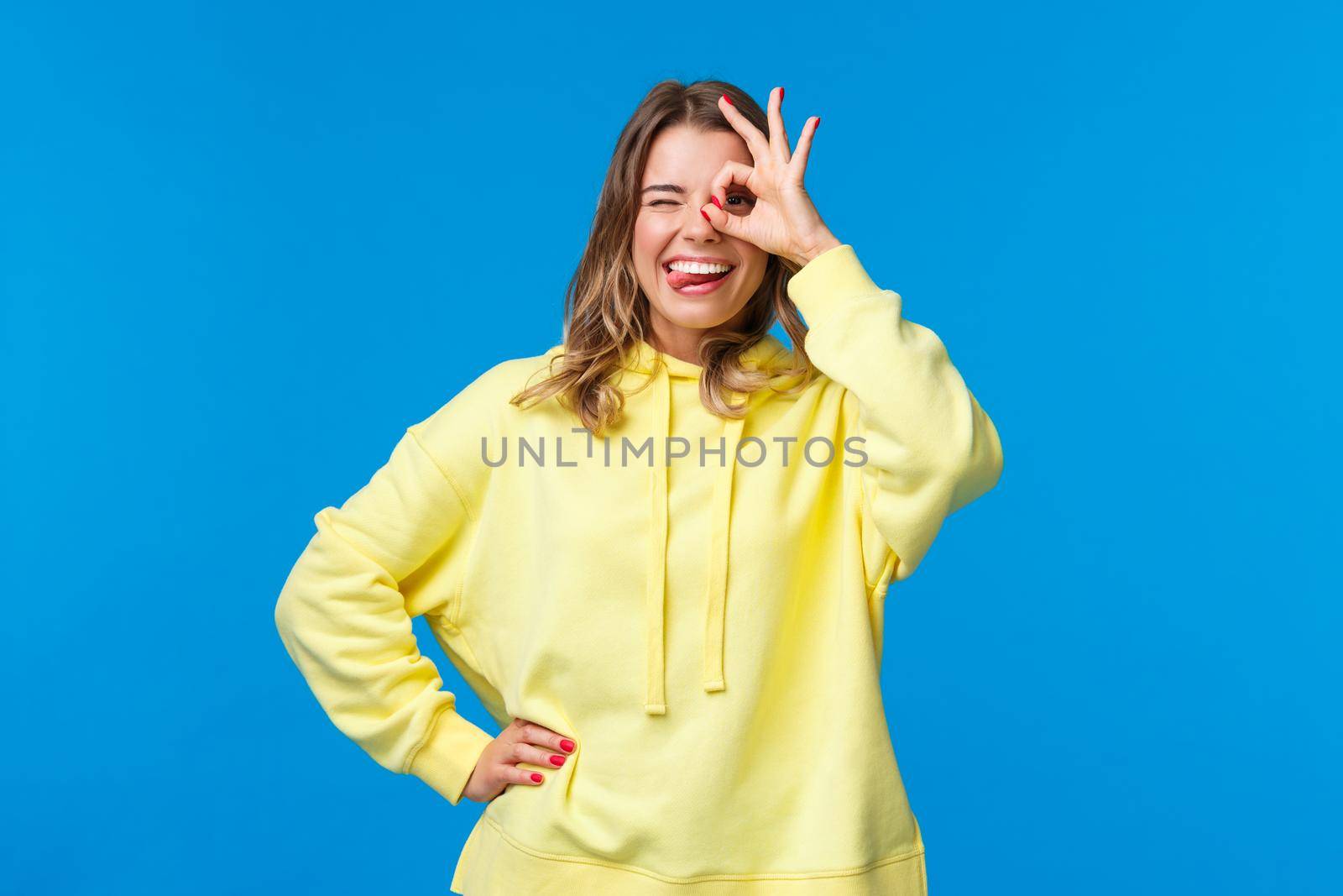 No problem, stay relaxed. Carefree and chill good-looking blond girl vibing, assure all okay nothing worry about, show tongue wink and look through okay gesture, blue background.