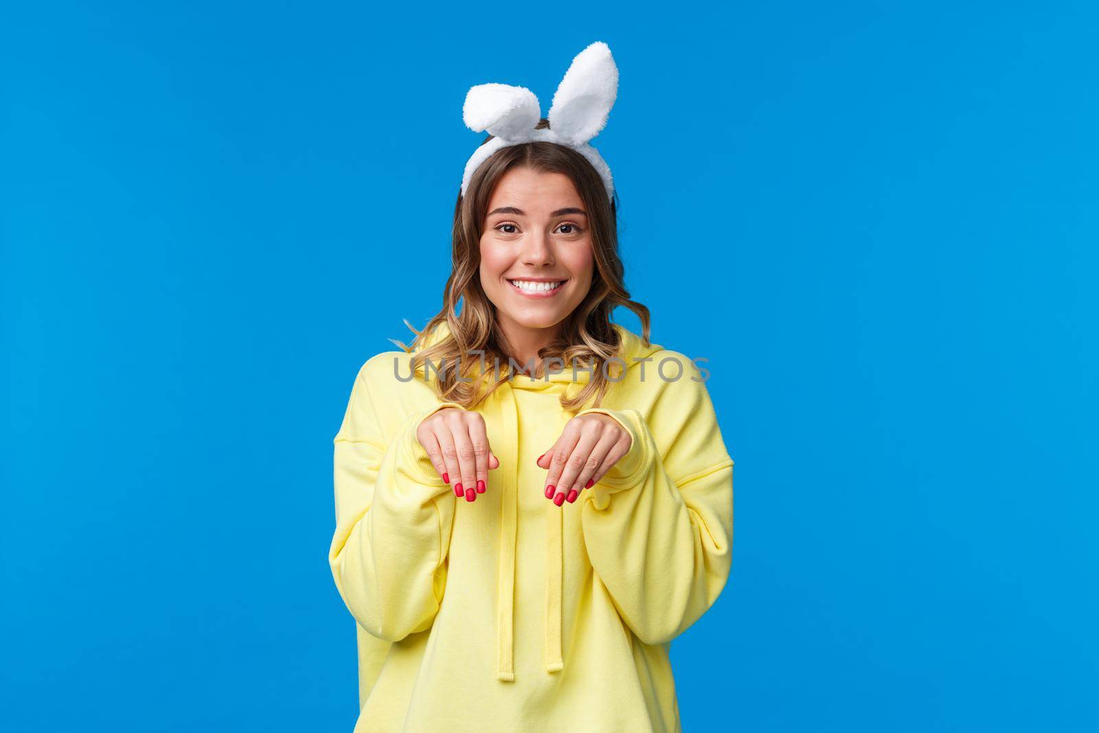 Holidays, traditions and celebration concept. Cute and silly smiling blond woman lovely looking at camera and mimicking rabbit with fake ears on head, standing blue background by Benzoix