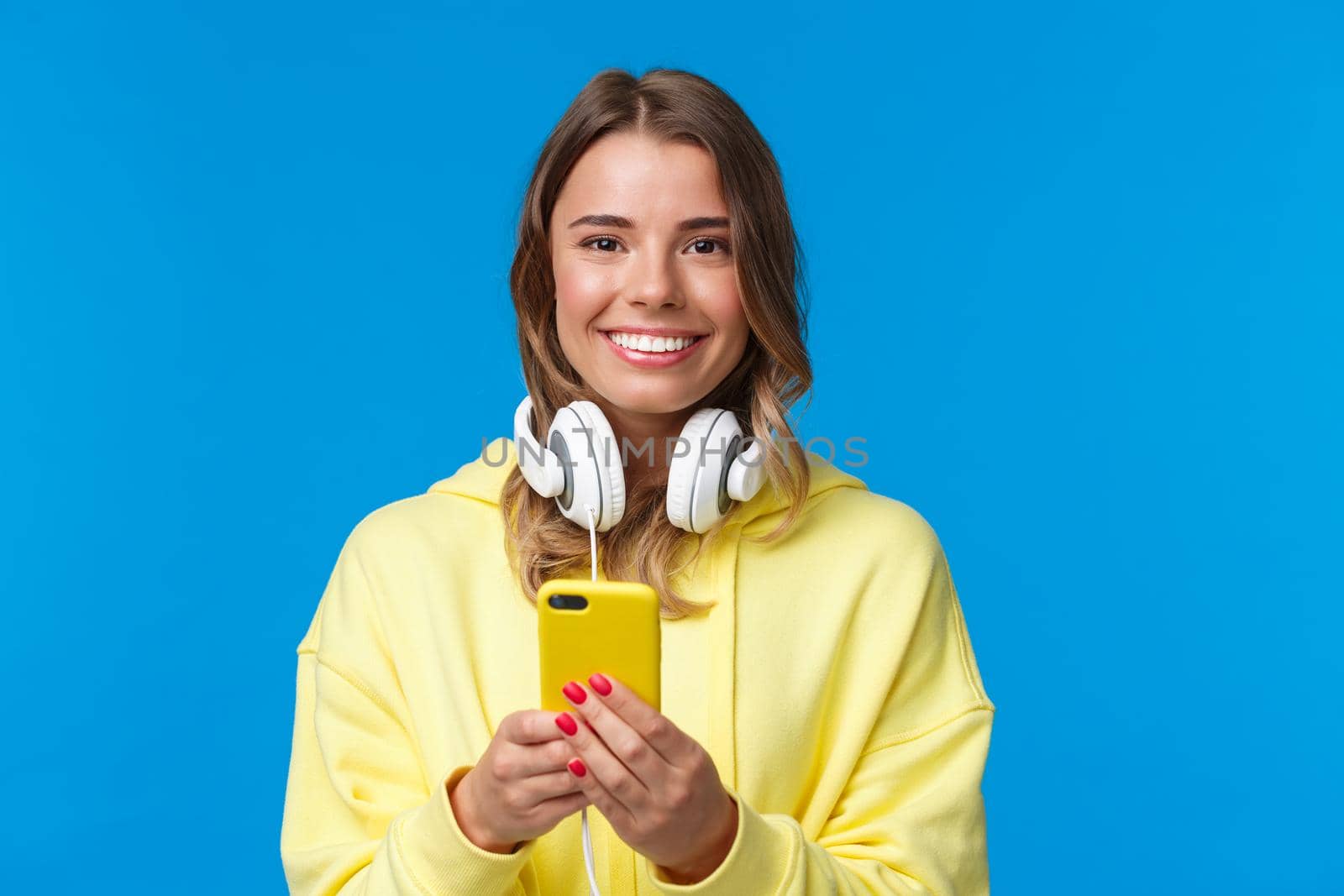 Close-up portrait of young blond european hipster girl with blond short hair and headphones over neck, holding smartphone, texting message, use mobile phone, blue background by Benzoix