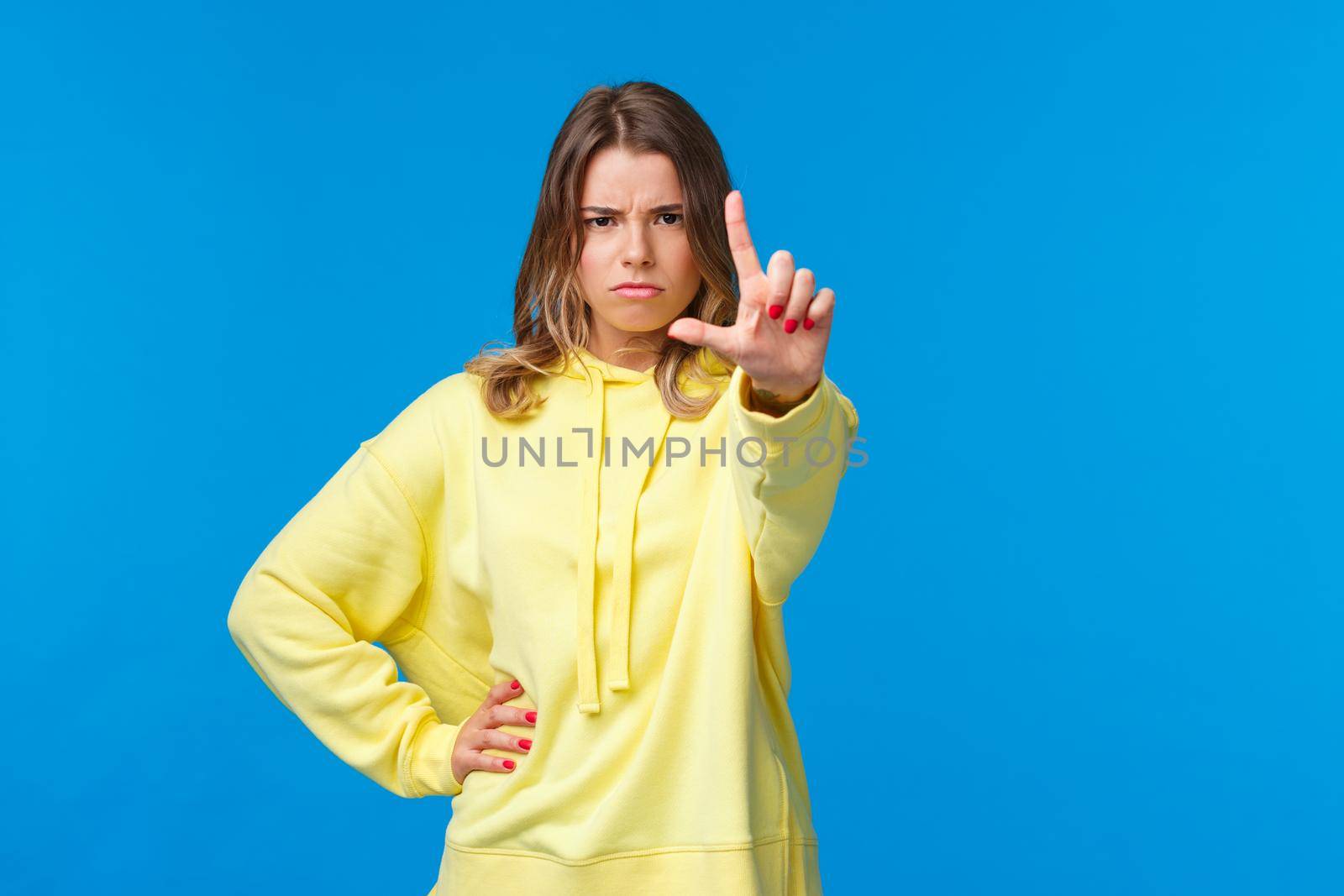 Stop right there. Serious-looking angry and bossy elder sister prohibit something, scolding kid as shaking finger with disapproval or disagreement, standing blue background.