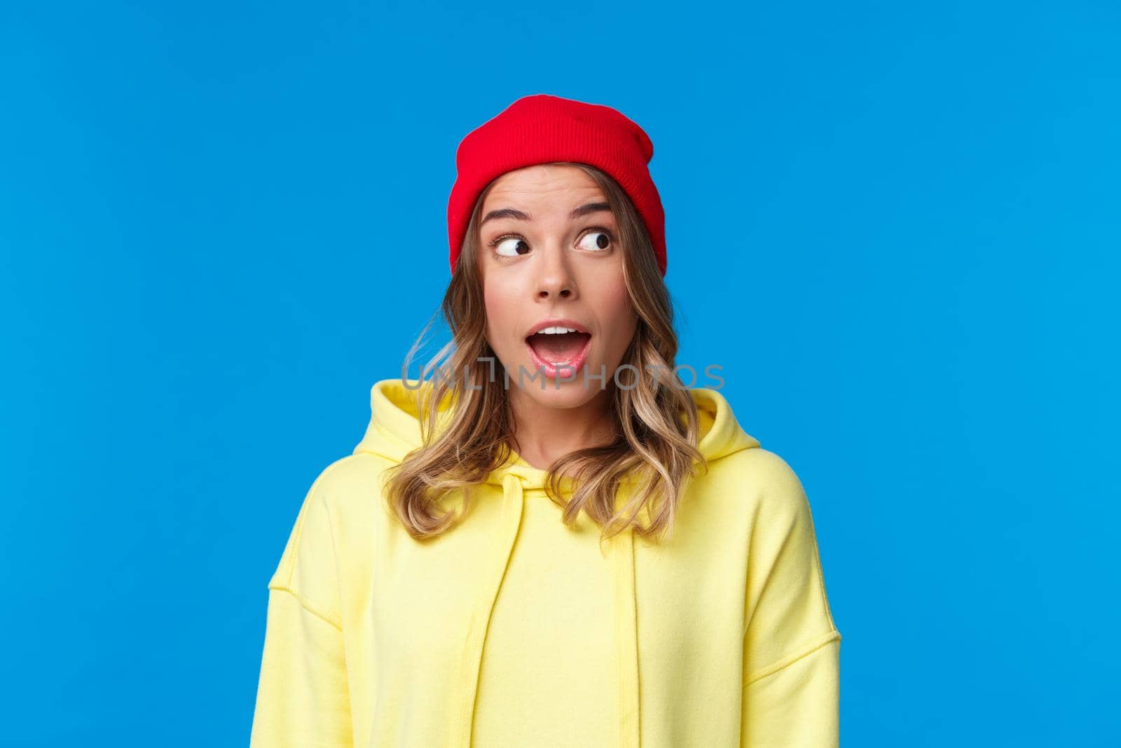 Excited good-looking caucasian female student in red beanie and yellow hoodie, look upper right corner and open mouth fascinated, hear interesting gossip, standing blue background by Benzoix