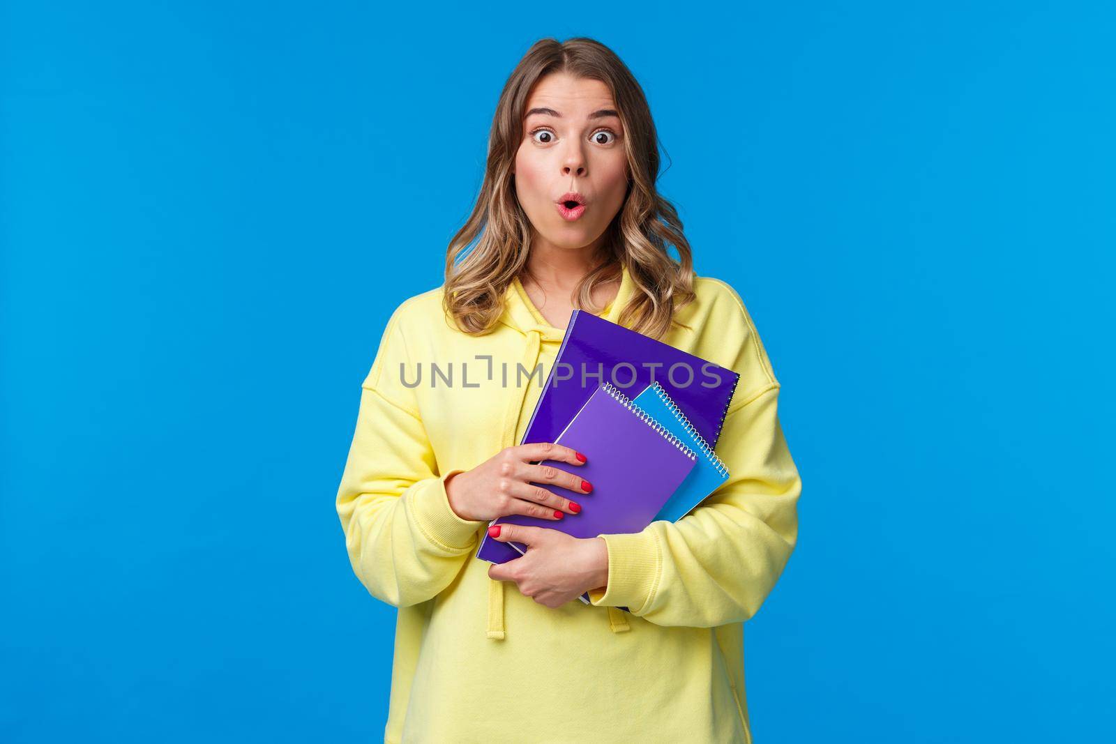 Waist-up portrait of surprised and wondered blonde caucasian female say wow and express amazement hear interesting news, gossiping in college, holding notebooks and essay, blue background by Benzoix