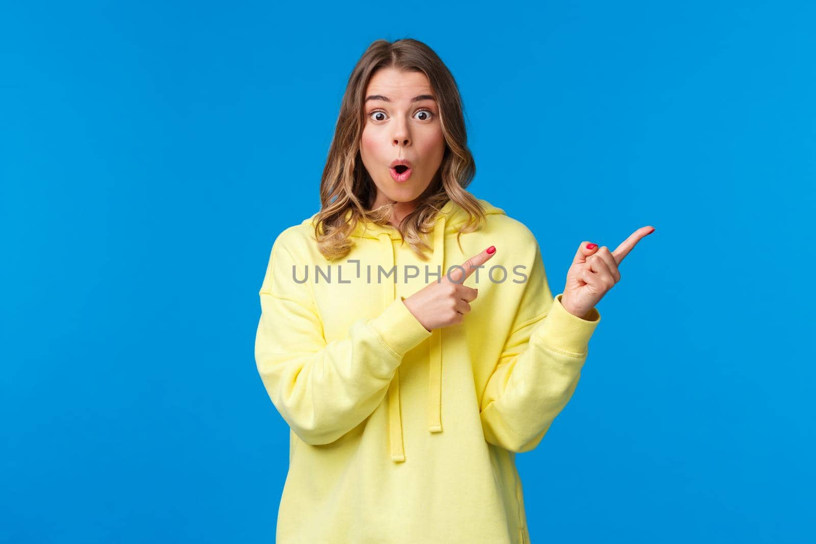 Have you seen it. Impressed and stunned blond caucasian female in yellow hoodie discuss recent events, gossips with friend, look at you and pointing right with dropped jaw, blue background.