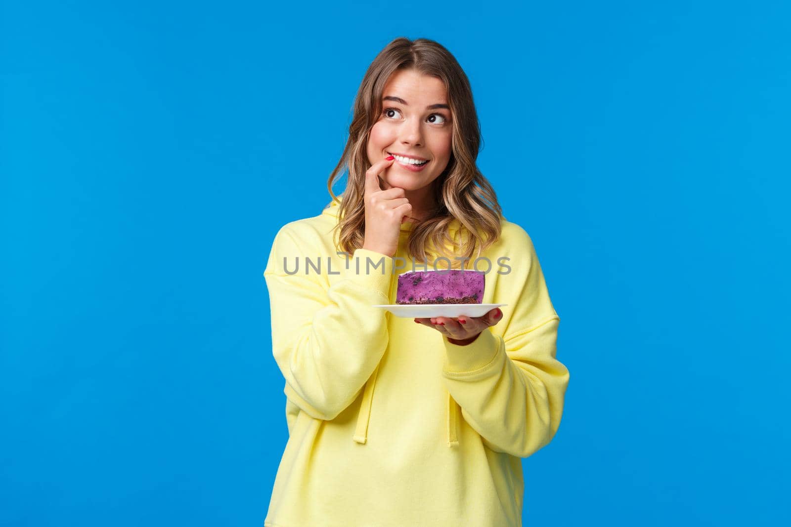 Celebration, party and lifestyle concept. Dreamy cute and silly blond girl thinking what to wish, biting finger and looking thoughtful up, smiling holding b-day cake, blue background.