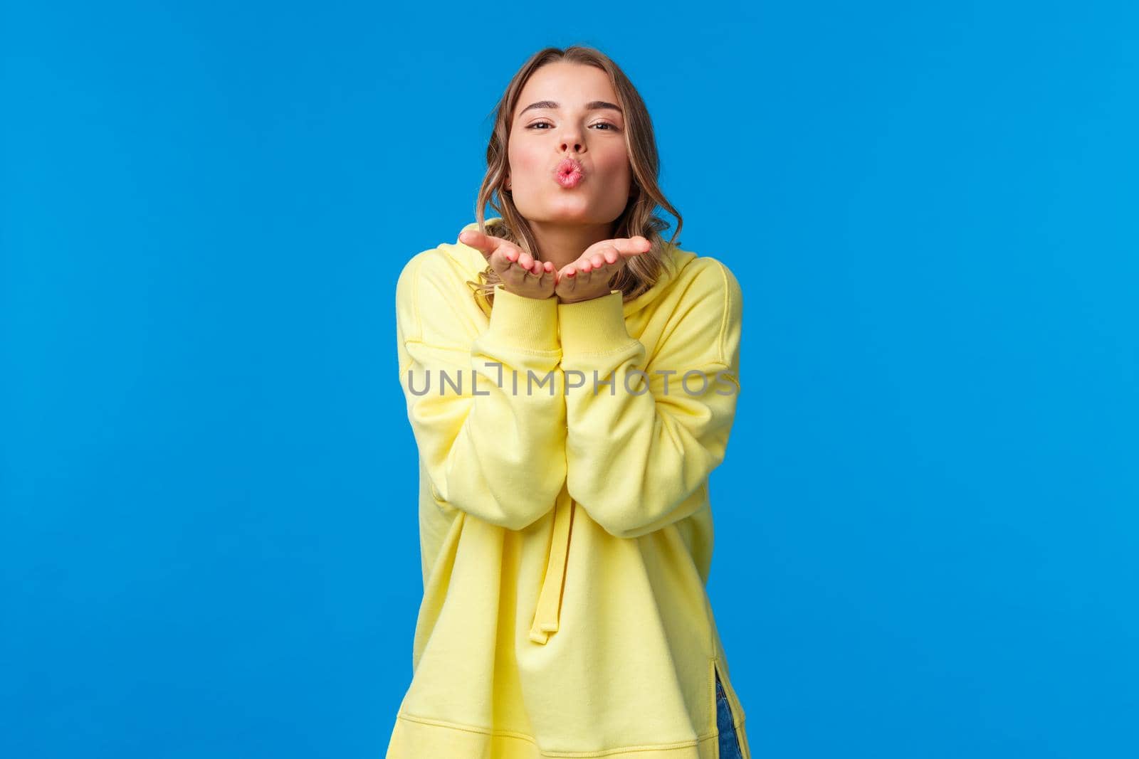Send you passionate kisses through camera. Tender and silly girlfriend with blond short hair holding palms near folded lips and make mwah gesture, standing blue background by Benzoix