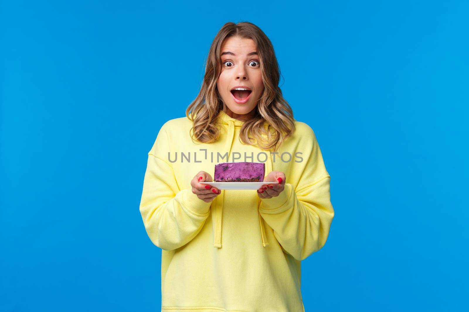 Celebration, party and lifestyle concept. Excited and happy cheerful blond girl on diet eating tasty fruit cake, looking surprised and joyful camera, standing blue background by Benzoix