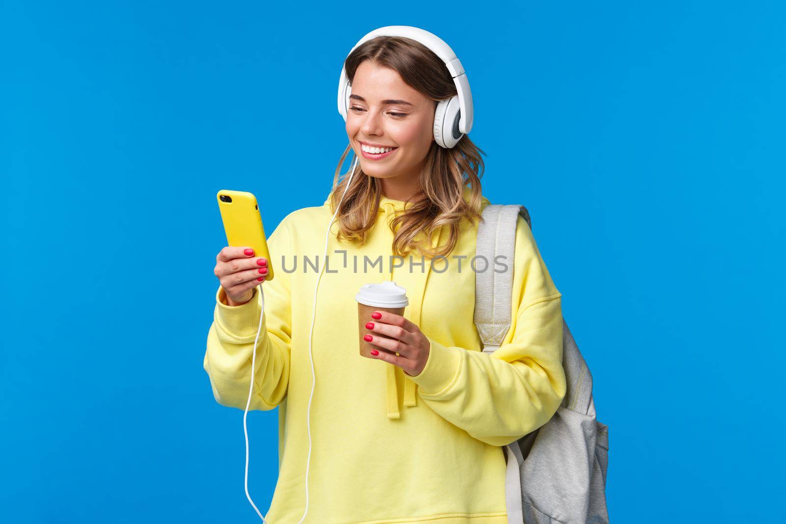 Carefree smiling blond woman in yellow hoodie listening music in headphones, picking song from playlist as drinking coffee on her way home from university, hold backpack, blue background by Benzoix