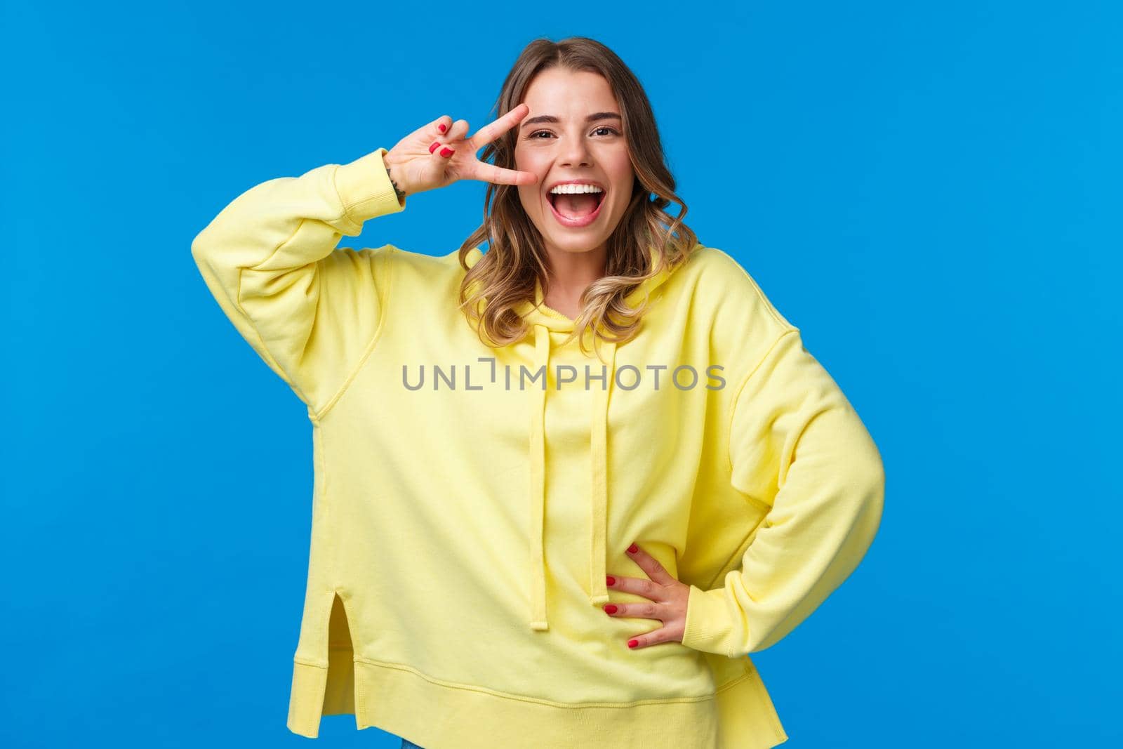 Portrait of happy optimistic laughing girl with blond short hair, smiling joyfully as show disco peace gesture near eye, posing for photo near blue background, having fun by Benzoix