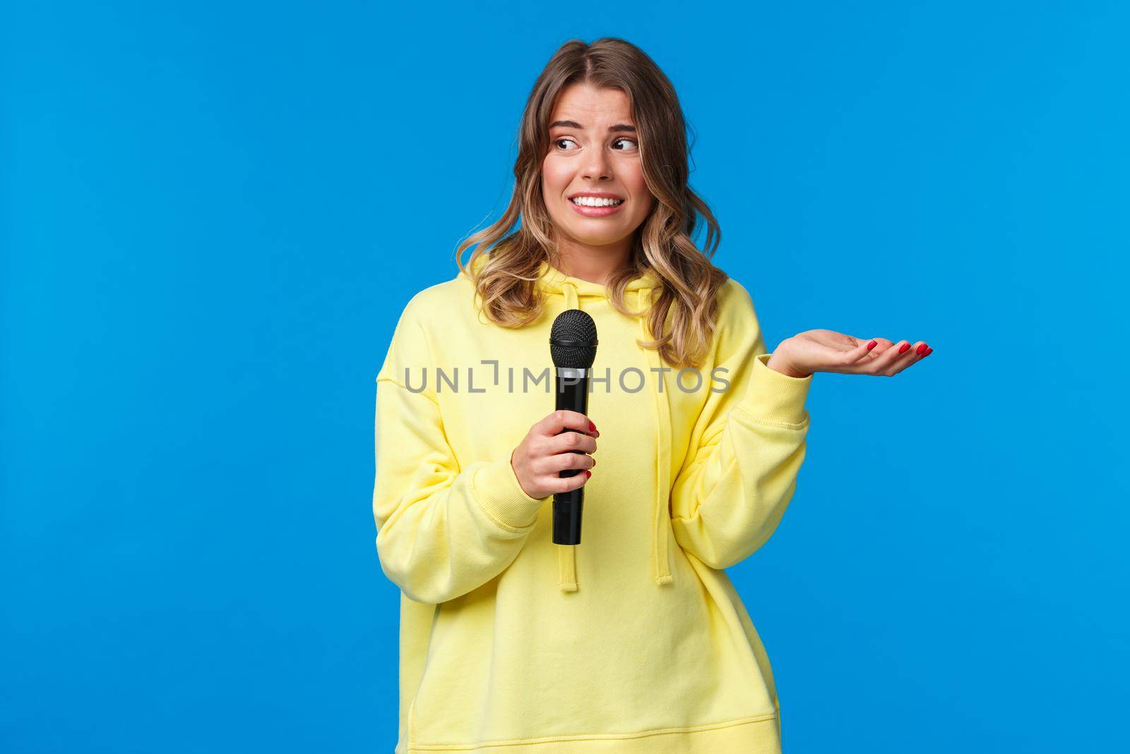 Awkward cute blond girl dont know lyrics as singing karaoke, looking at friend nervous smile, embarrassed sing in fron of people, hold microphone, standing blue background by Benzoix