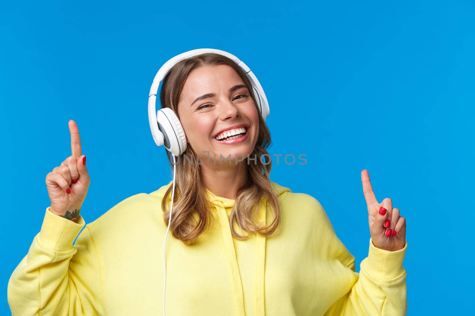 Music, lifestyle and youth concept. Close-up portrait of joyful smiling blond girl listening songs in headphones, dancing and move fingers rhythm, standing happy over blue background by Benzoix