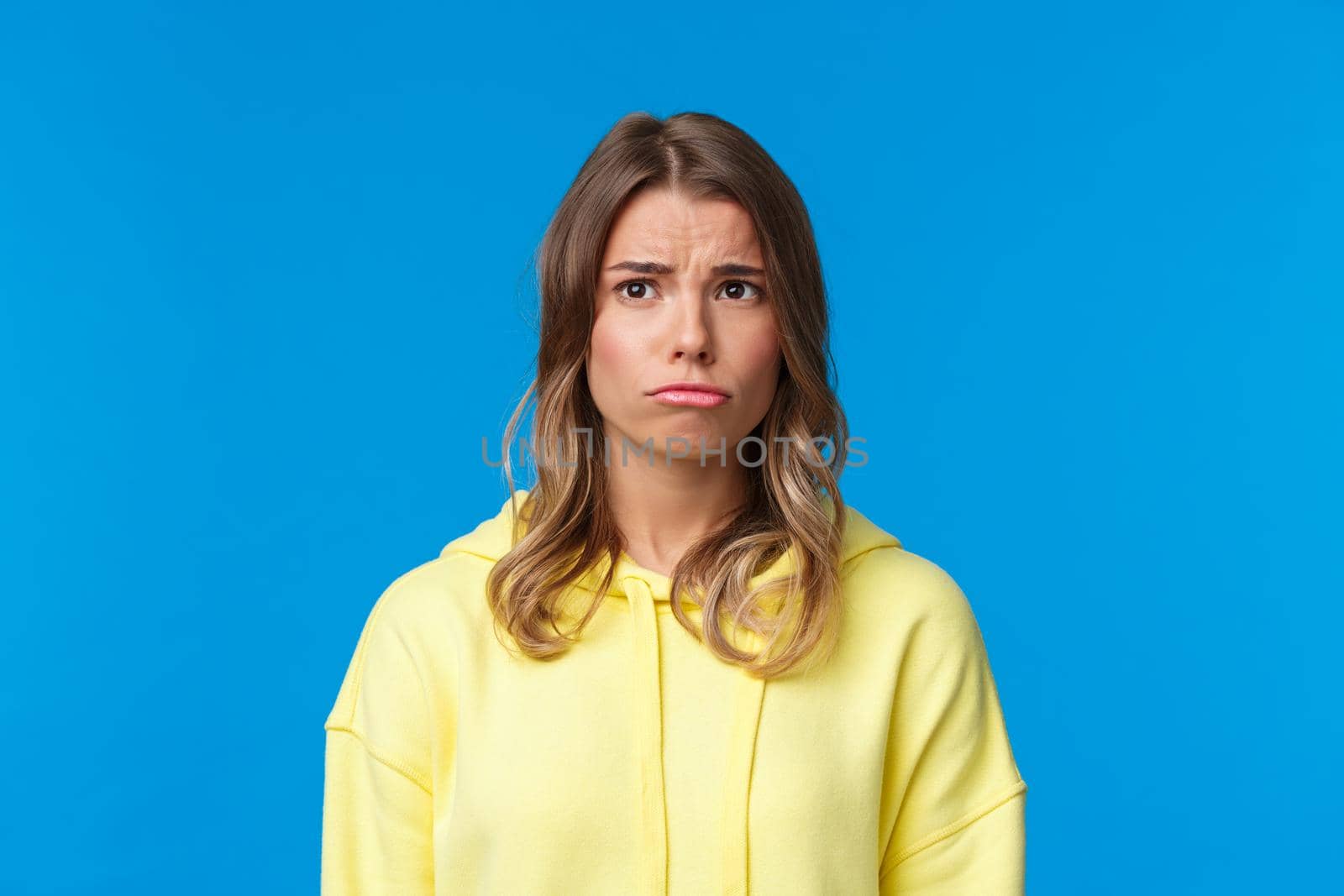 Close-up portrait gloomy sad cute jealous blond girl in yellow hoodie, pouting looking with regret upper left corner, feeling moody and uneasy, standing blue background unhappy by Benzoix