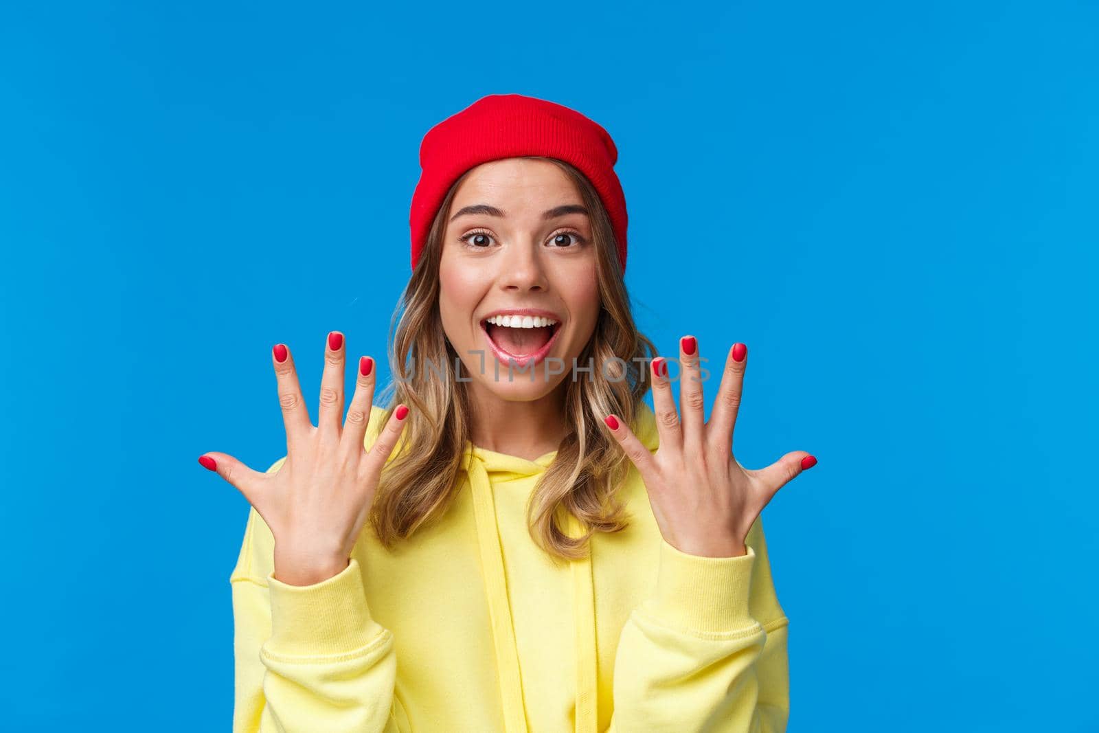 Portrait of amused good-looking blonde hipster girl smiling joyfully, explain something showing number ten, tenth with hands and look camera upbeat, order dozen, stand blue background.