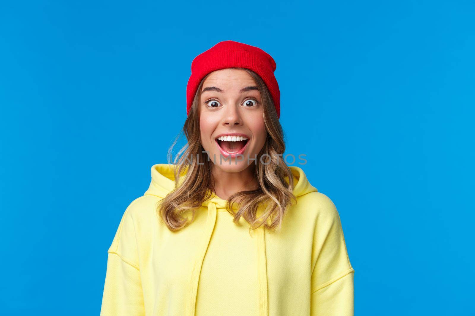 Close-up portrait of excited and intrigued young hipster girl in red beanie and yellow hoodie, gasping amazed look camera wondered and entertained, standing blue background.