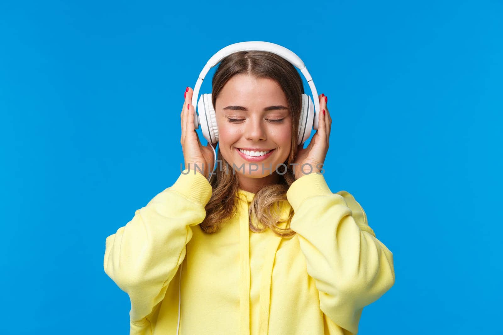 Music, lifestyle and youth concept. Close-up portrait of dreamy happy and relaxed young blond european girl close eyes and enjoying fine sound of song in headphones, dreaming, blue background.